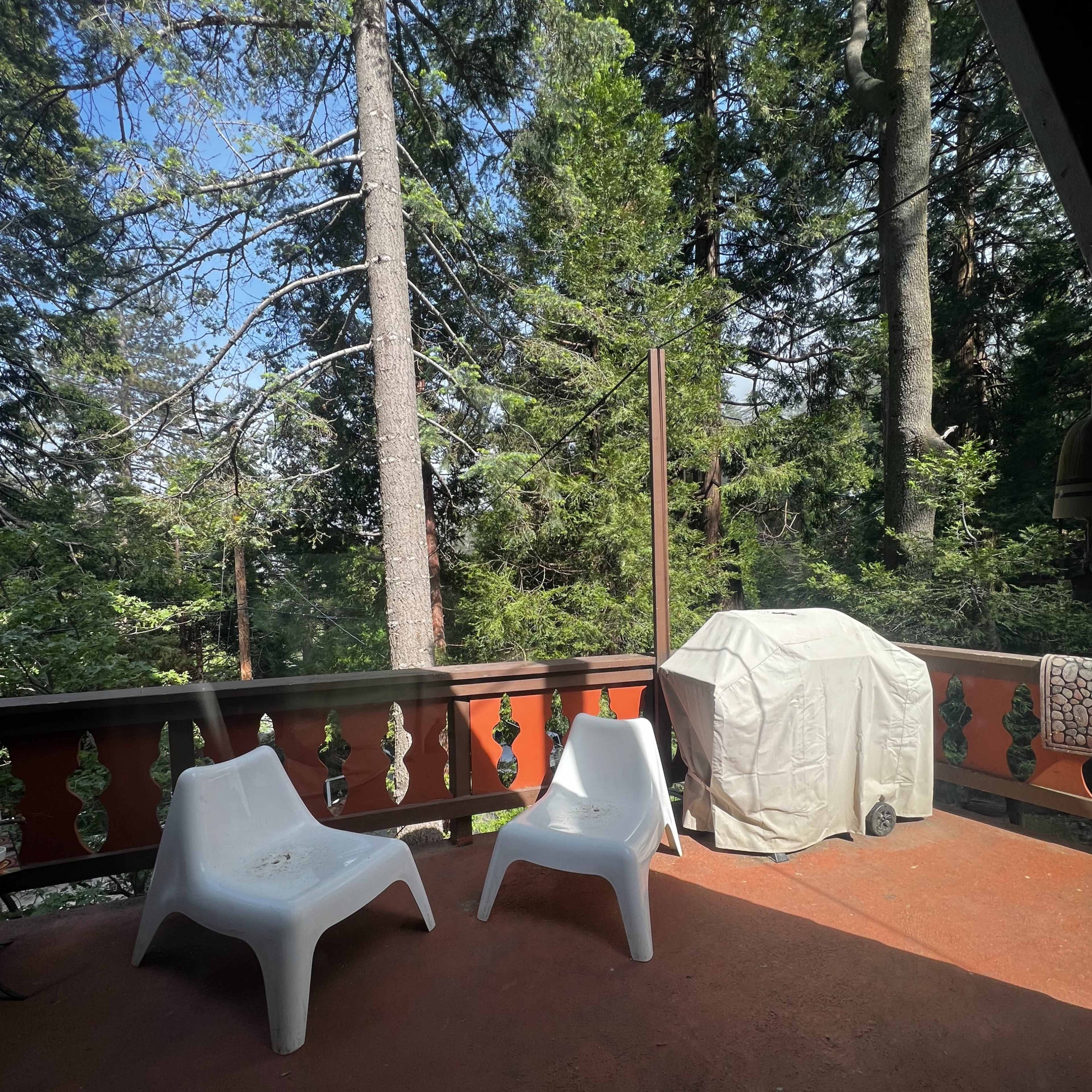 A small outdoor deck features two white plastic chairs and a covered grill, surrounded by tall trees.