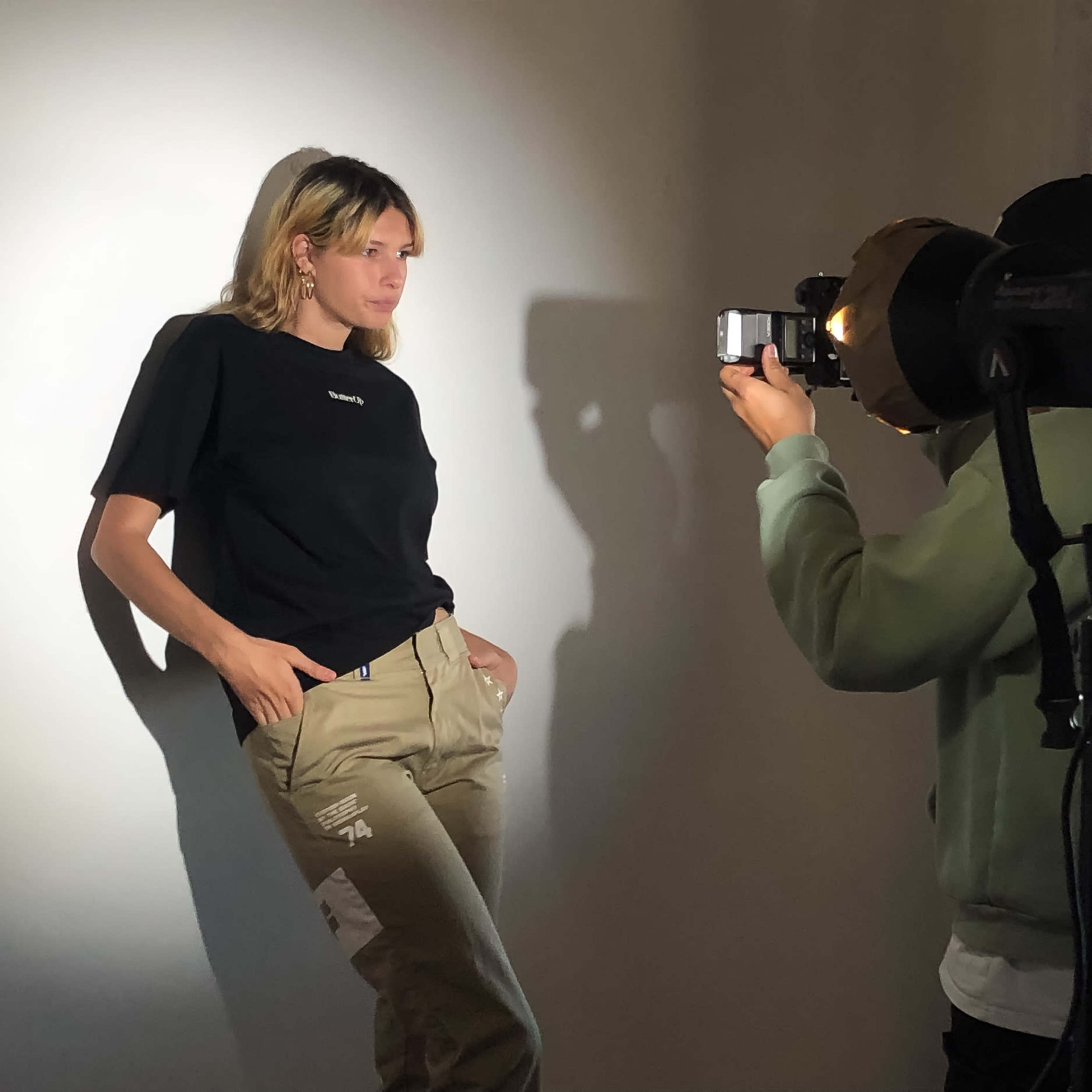 A woman poses against a wall while a man photographs her using a camera with a light source.