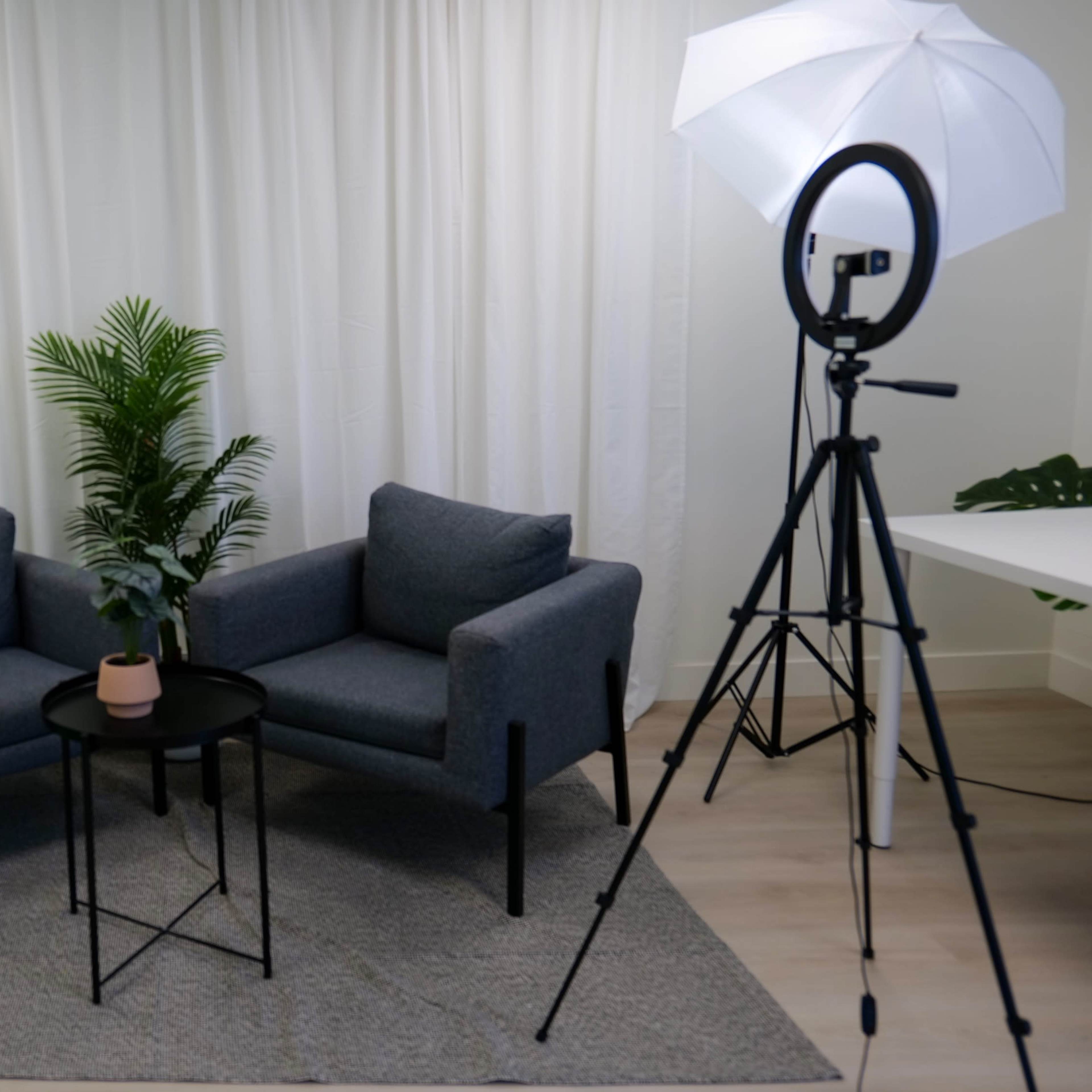 A simple studio setup with two grey chairs, a small black table, a potted plant, and lighting equipment, all against a backdrop of white curtains and a green screen.