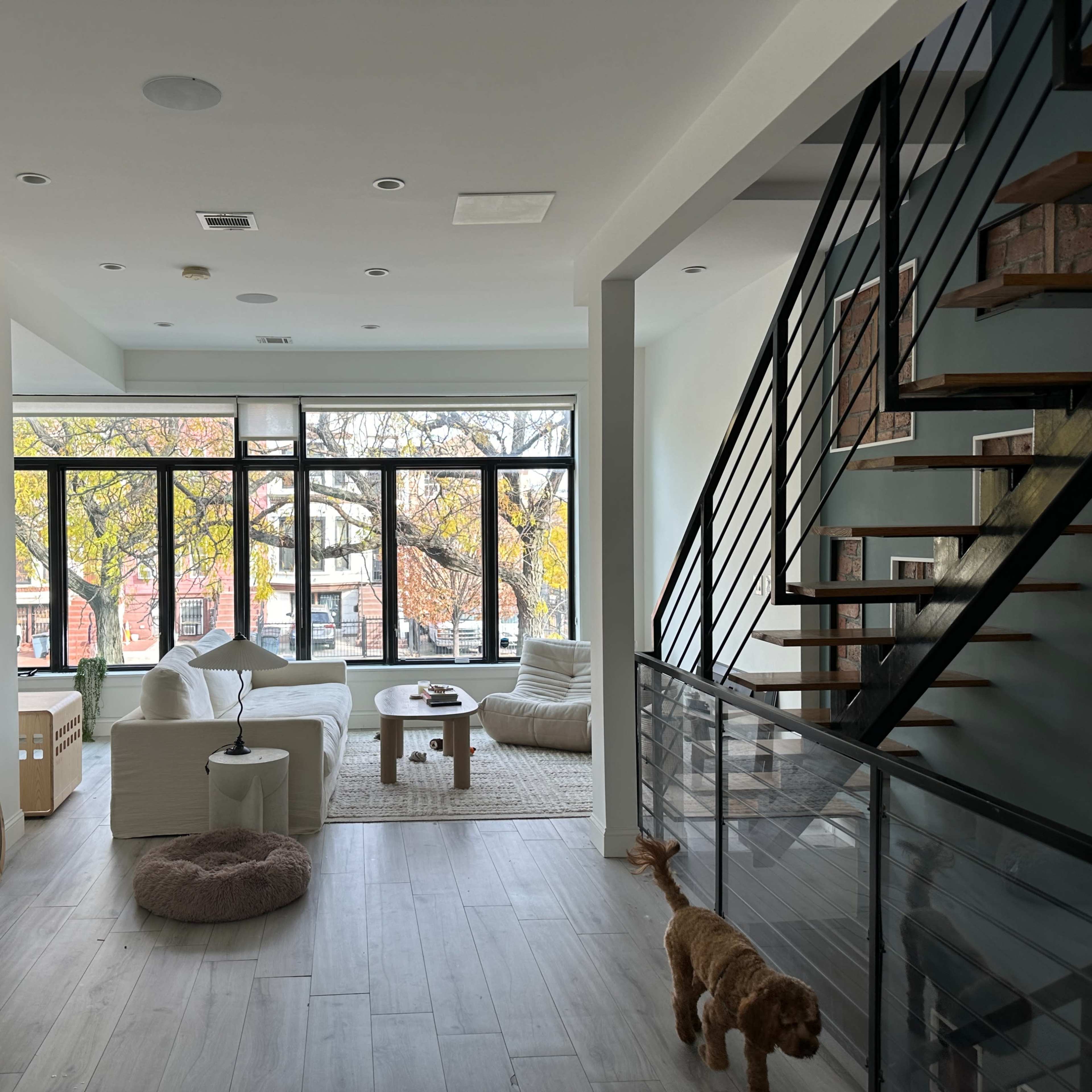 A modern, open-concept living area features a white seating arrangement, a large window overlooking trees, and a staircase with wooden steps.