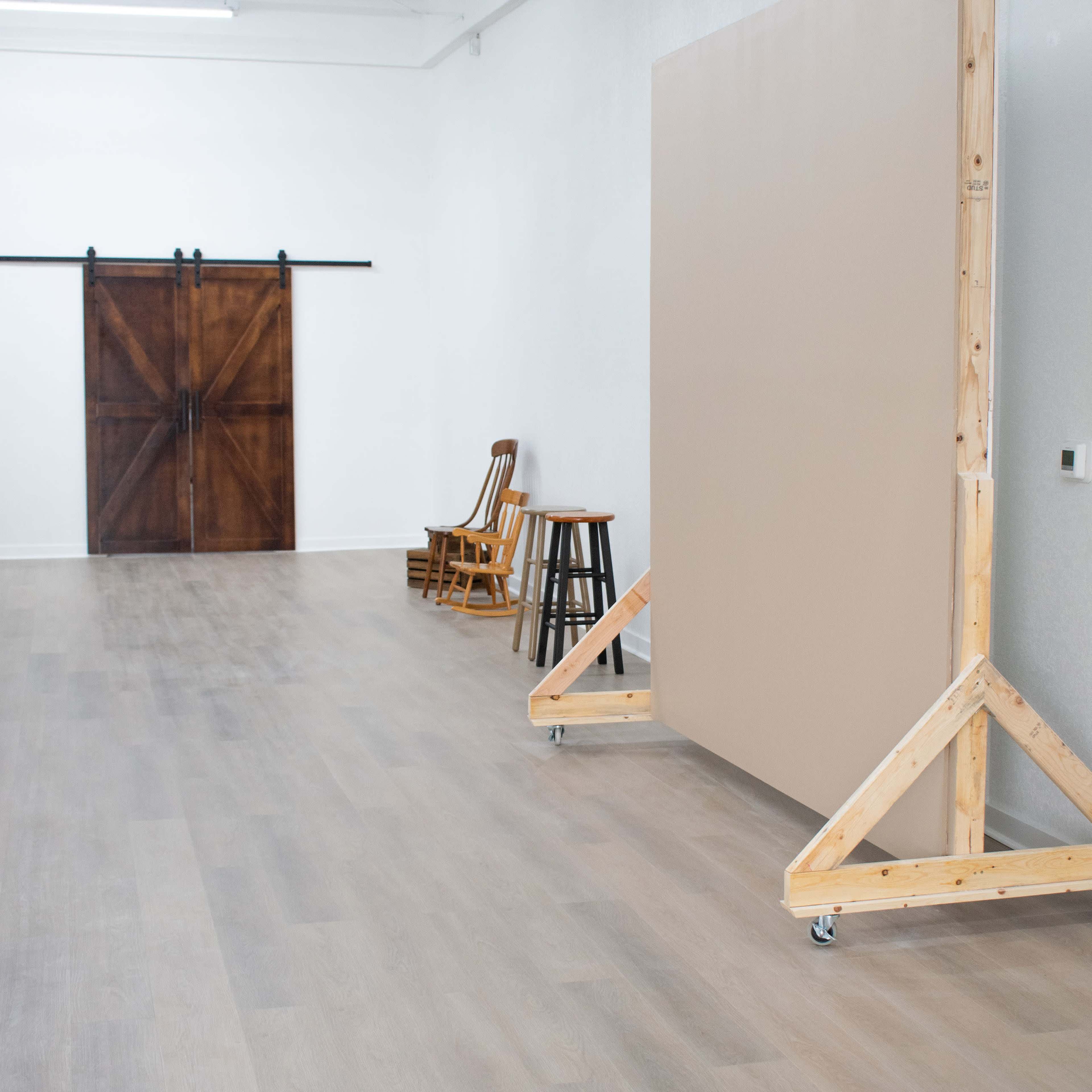 A spacious interior with wooden flooring, featuring a large, movable beige panel, three wooden chairs, and a set of sliding barn doors in the background.