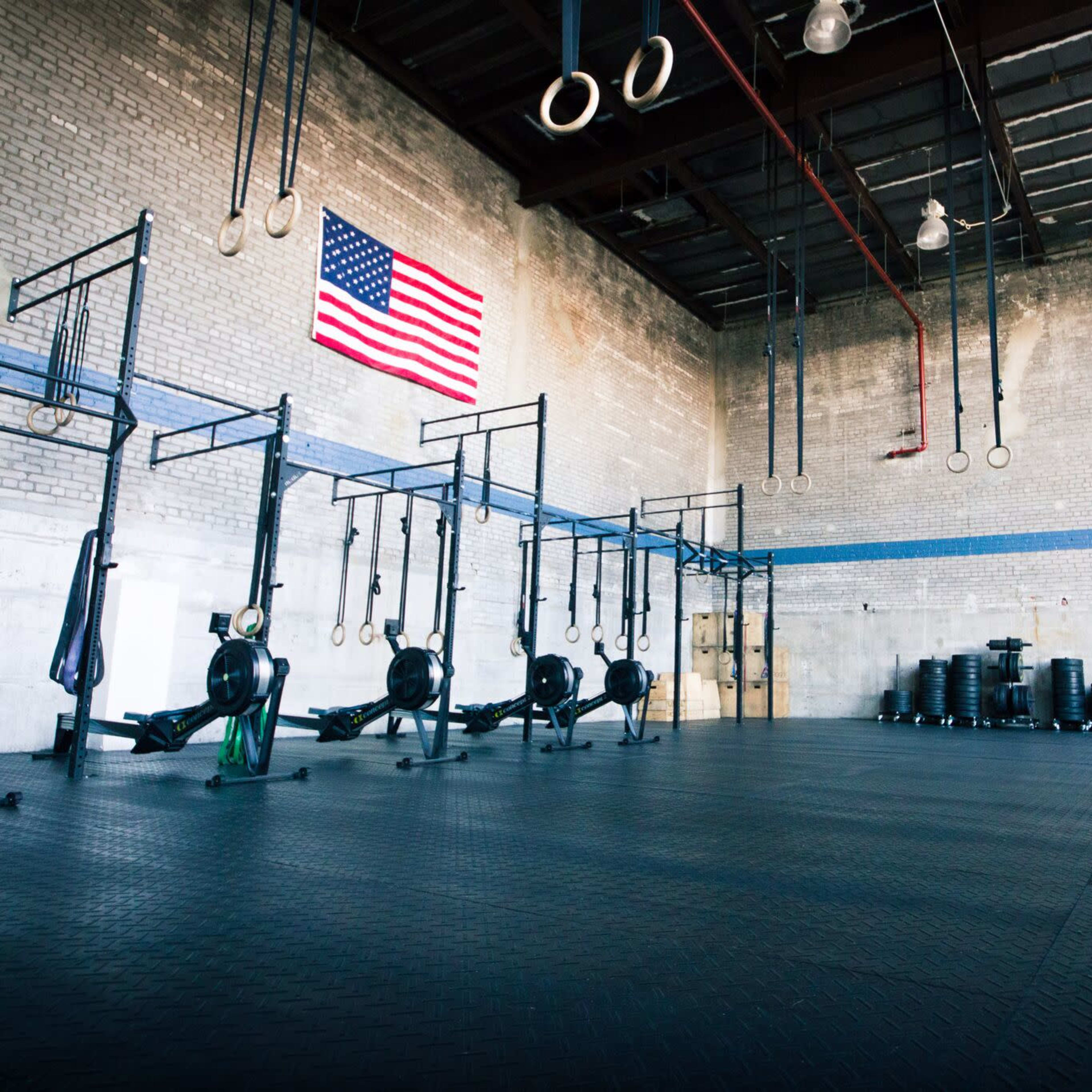 A spacious gym with rowing machines, gymnastic rings, and a large American flag on the wall.