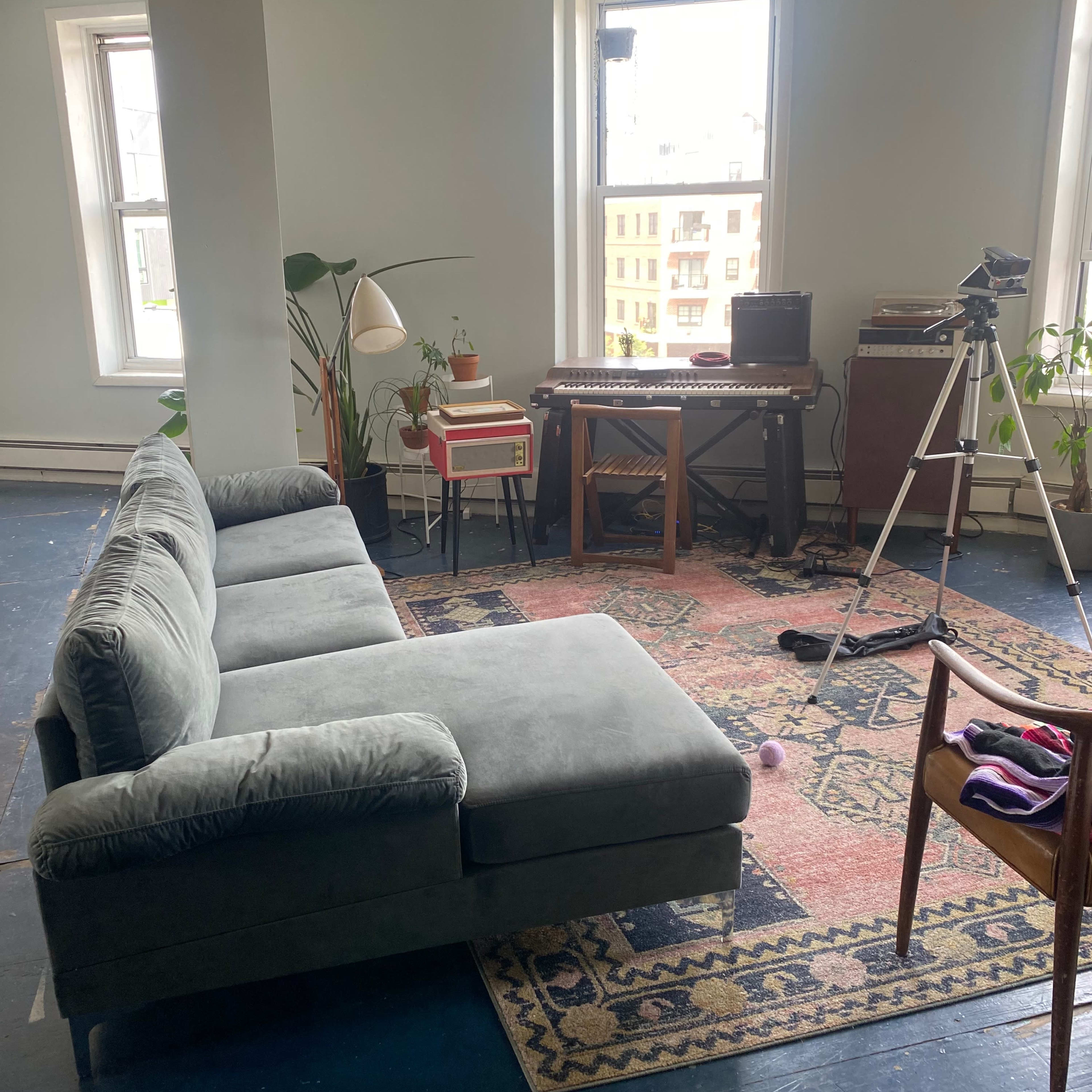 A gray sectional sofa is positioned on a rug in a light-filled room, with a plant, a small red side table, a keyboard on a wooden stand, and a tripod set up nearby.
