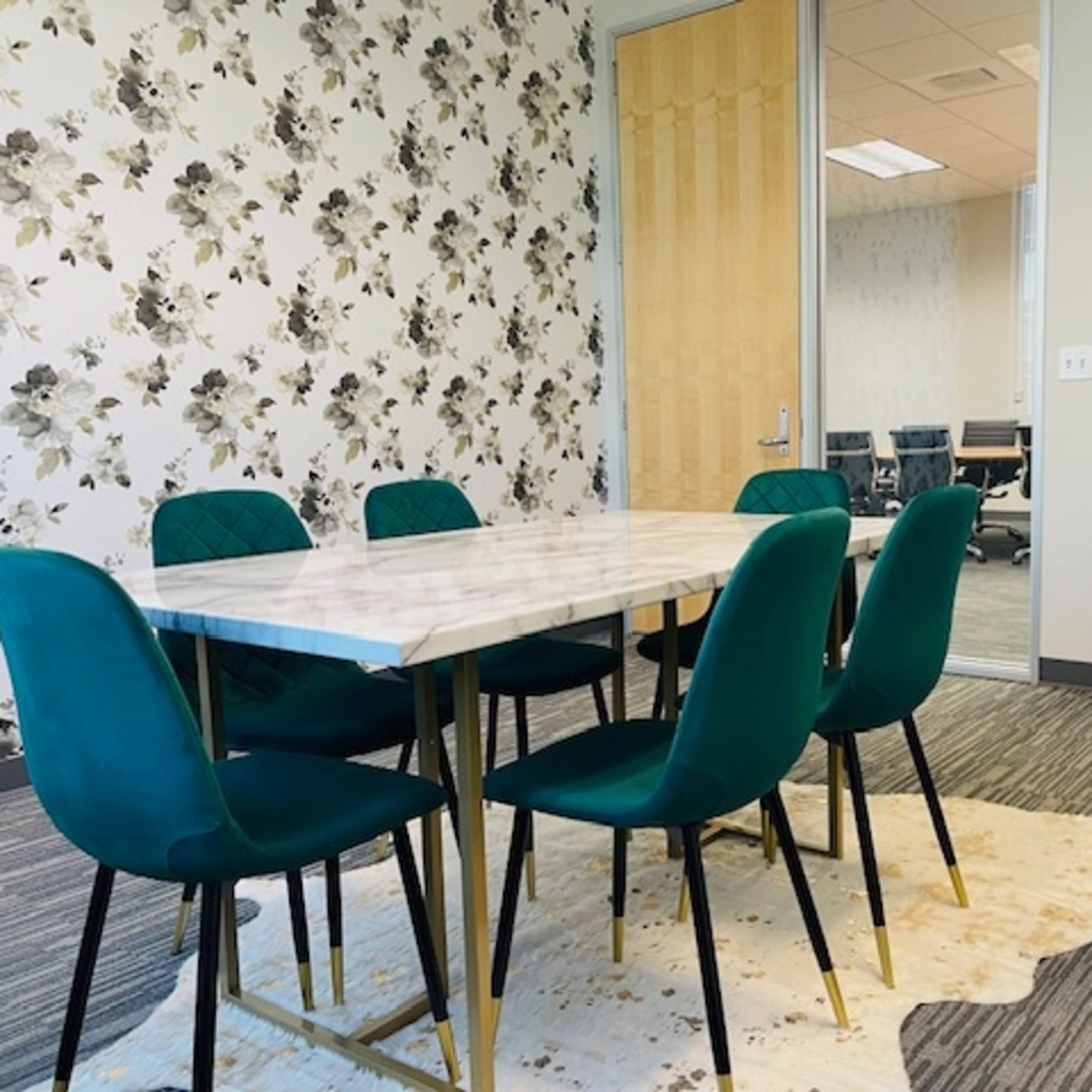 A conference room features a marble-top table surrounded by six green chairs on a patterned carpet, with a TV mounted on the wall and a glass-walled meeting area visible in the background.