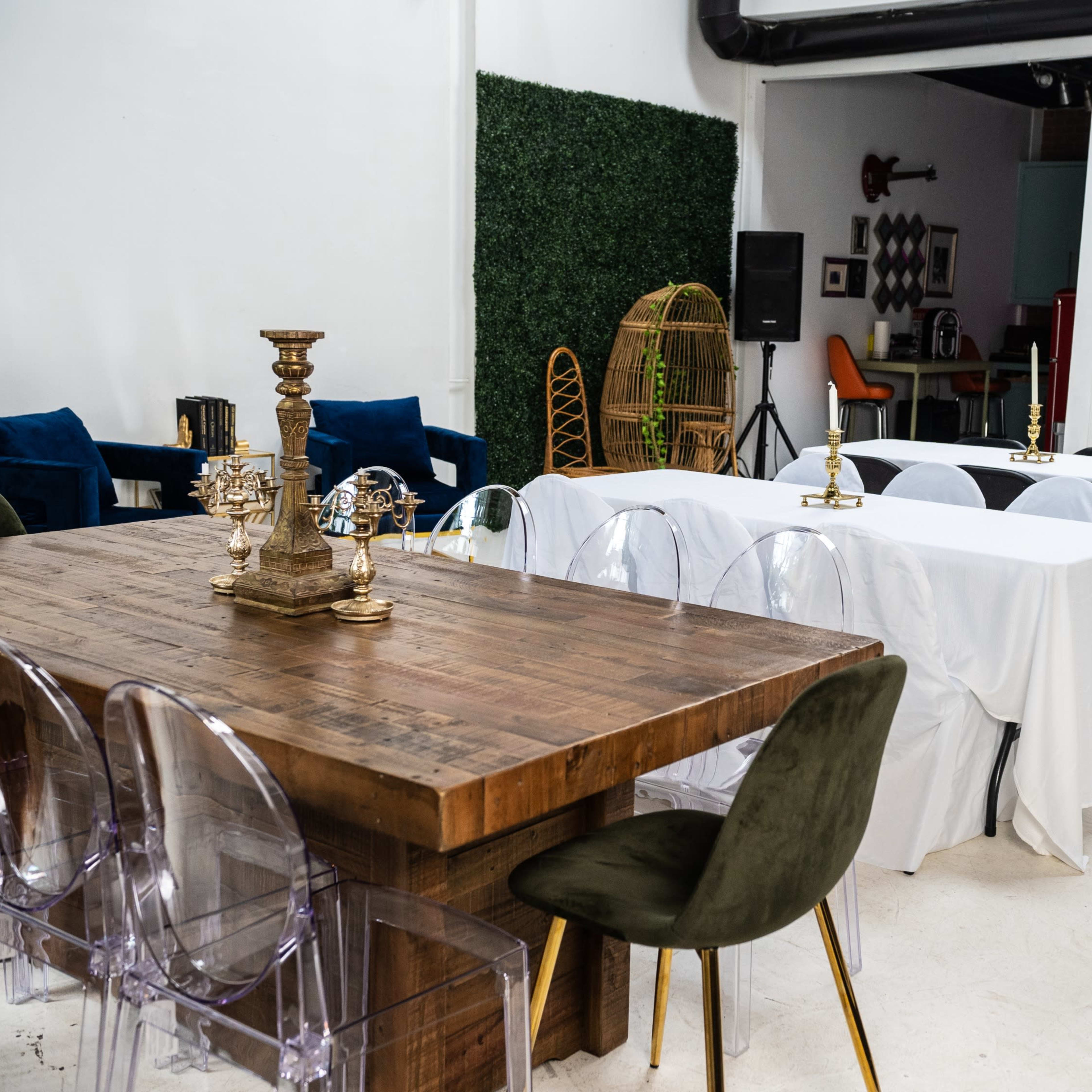 A modern event space featuring a large wooden table surrounded by transparent chairs and several covered tables in a well-lit room with greenery decorations.