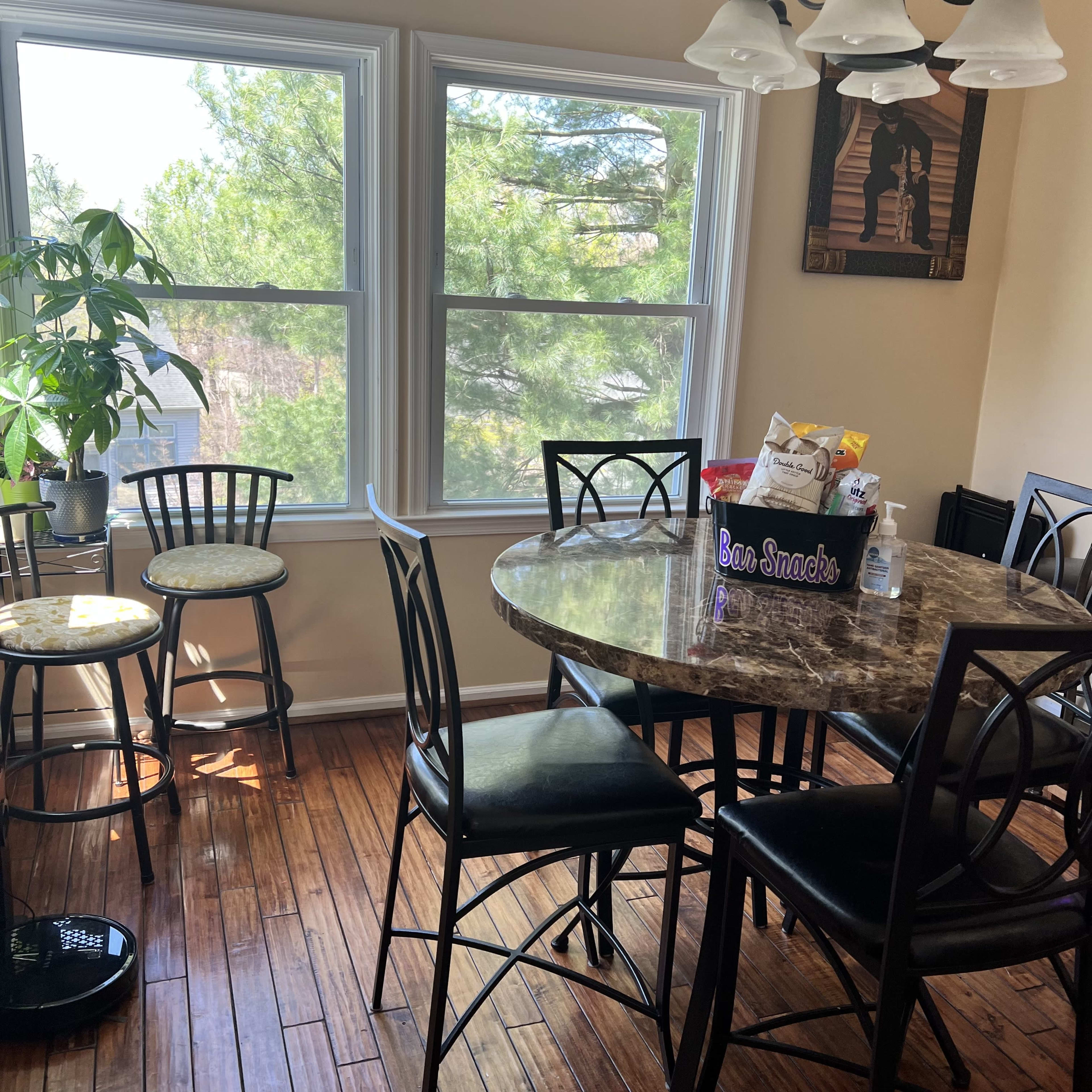 A round marble dining table with black chairs sits in a brightly lit room, featuring large windows that overlook greenery outside.