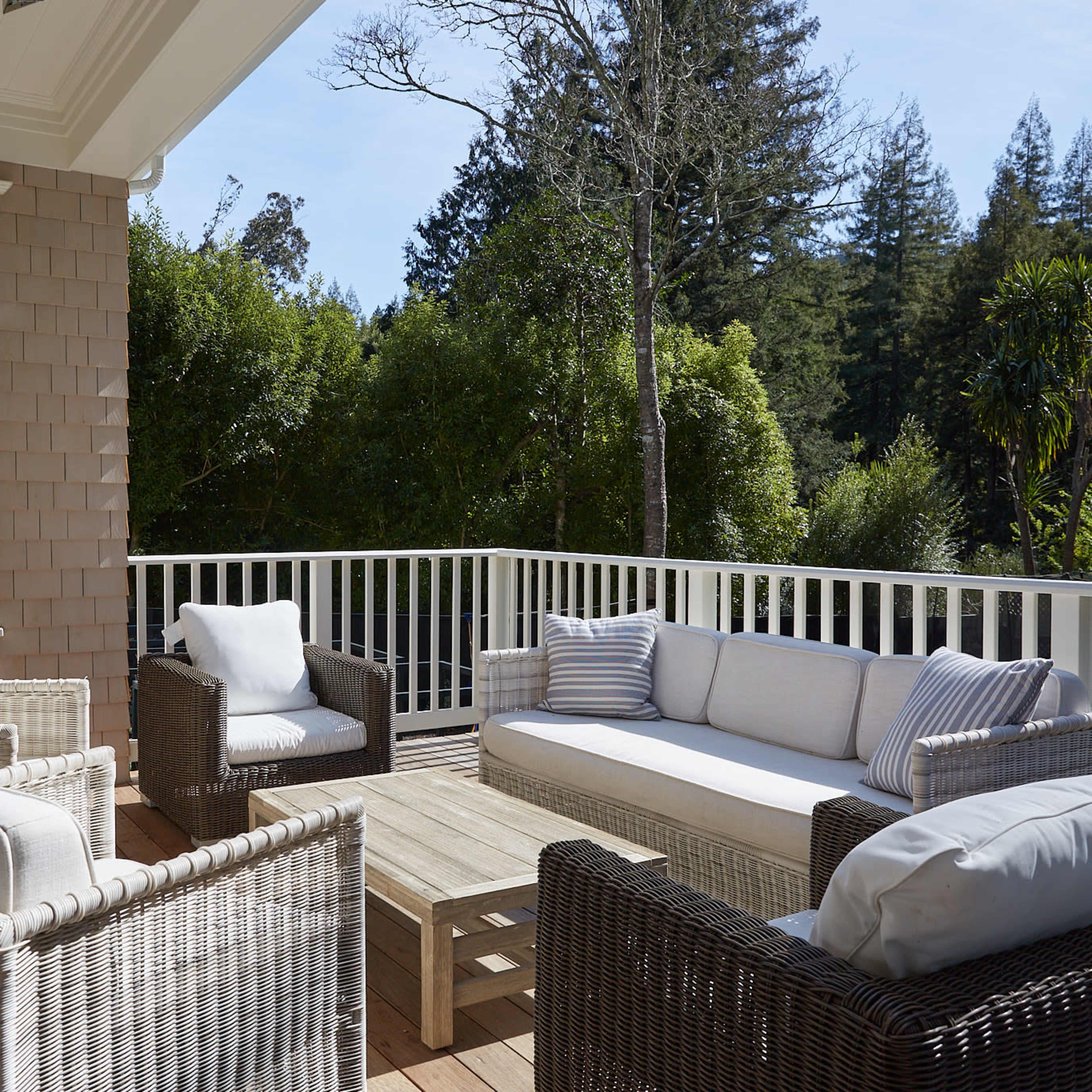 A covered porch features a seating arrangement of wicker furniture with light-colored cushions and a wooden coffee table, surrounded by greenery.