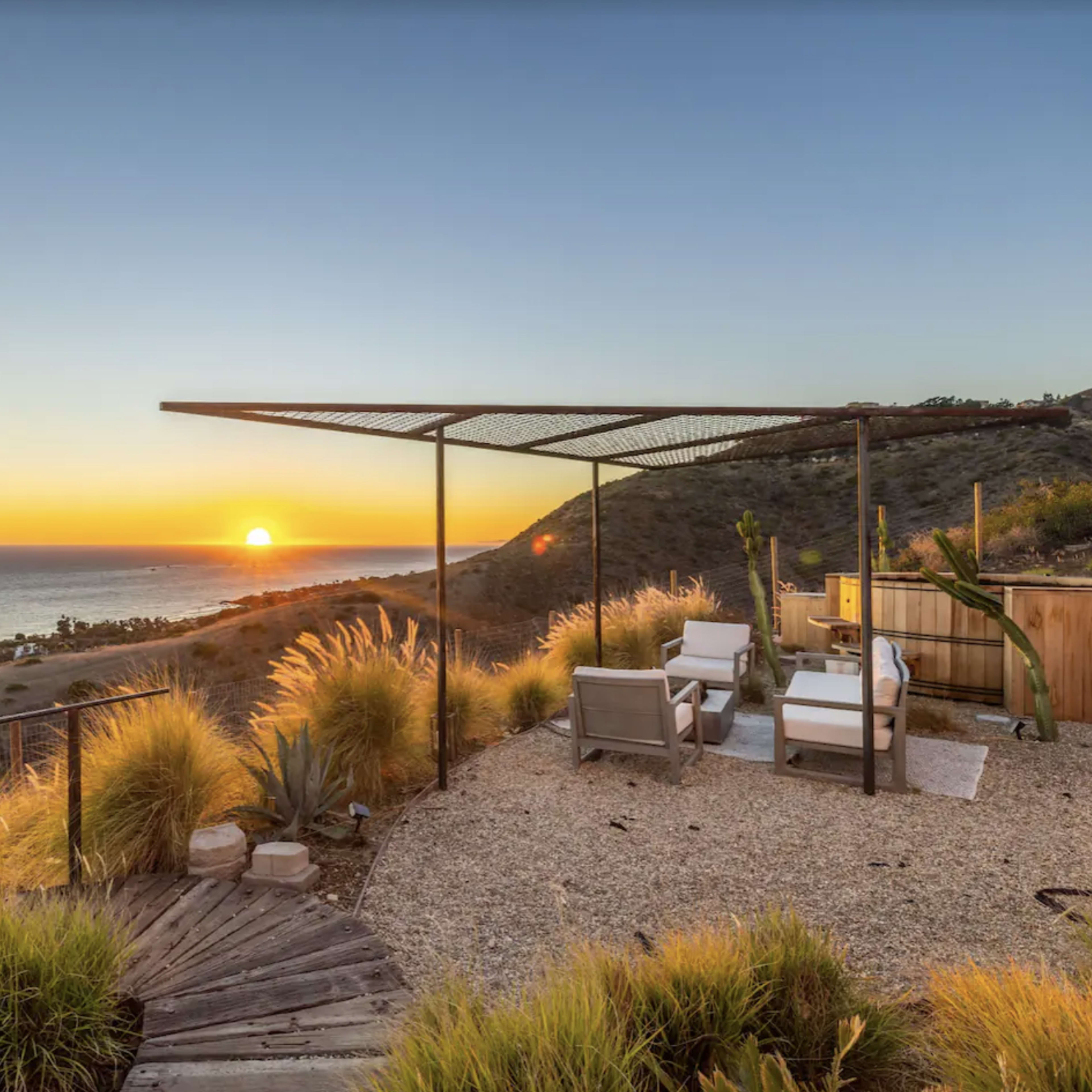 A hillside patio with chairs and a hot tub overlooking the ocean at sunset.