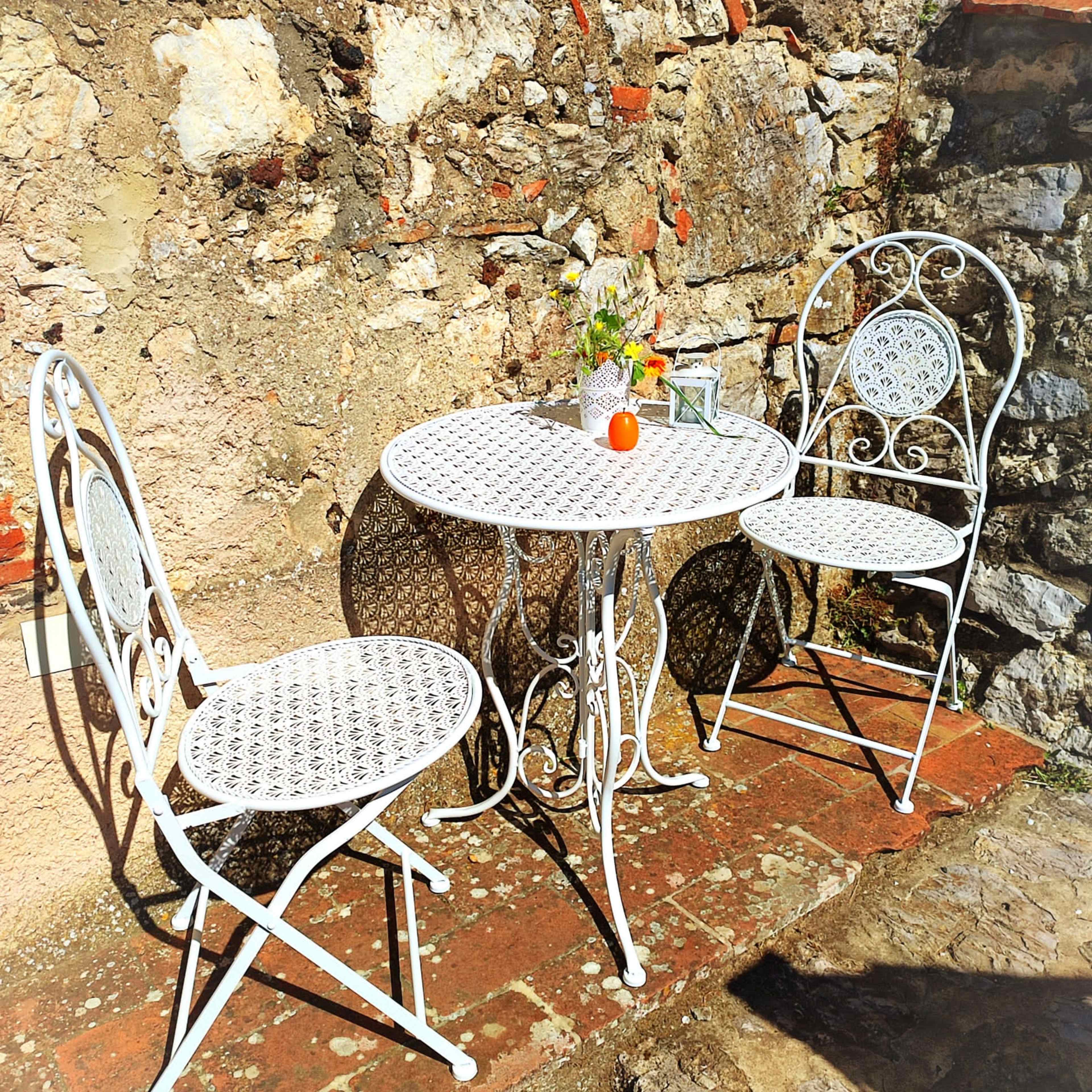 A small, round table with two matching wrought iron chairs is set on a stone patio, accompanied by a vase of flowers and a decorative orange.