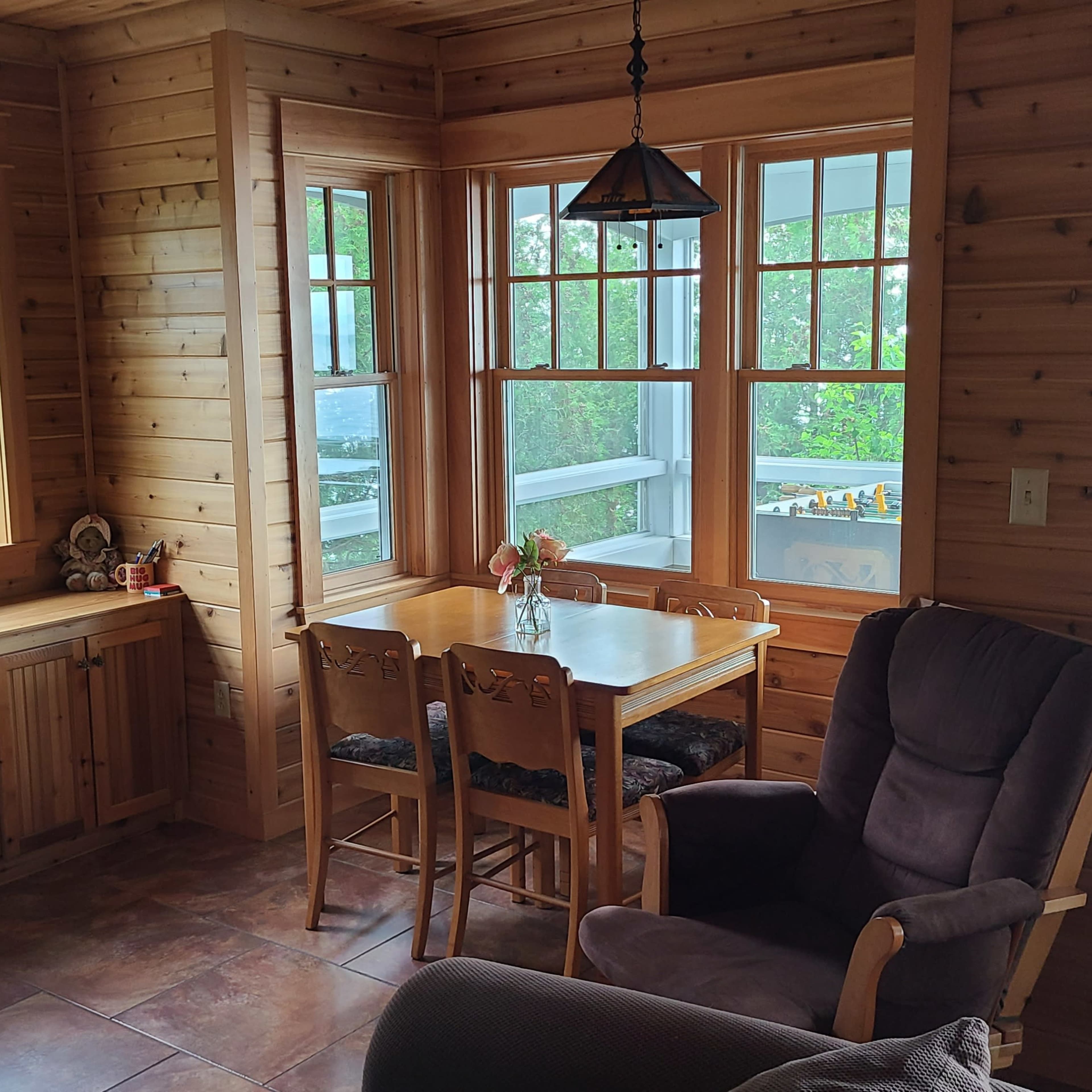 A cozy wooden room with a dining table, chairs, and a stone fireplace, illuminated by natural light from large windows.