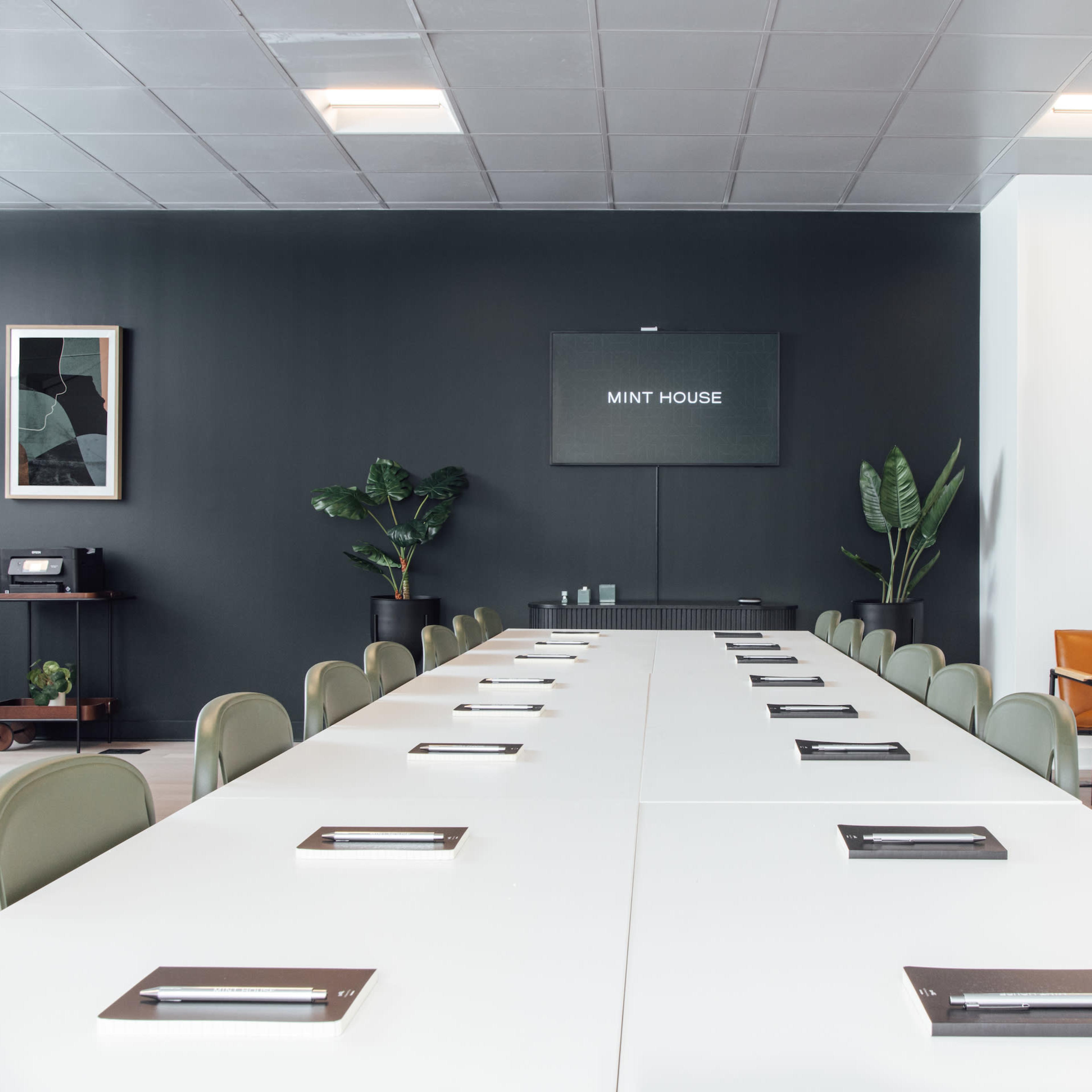 A modern conference room features a long white table with notepads, surrounded by green chairs and set against a dark wall displaying a sign that reads "MINT HOUSE."
