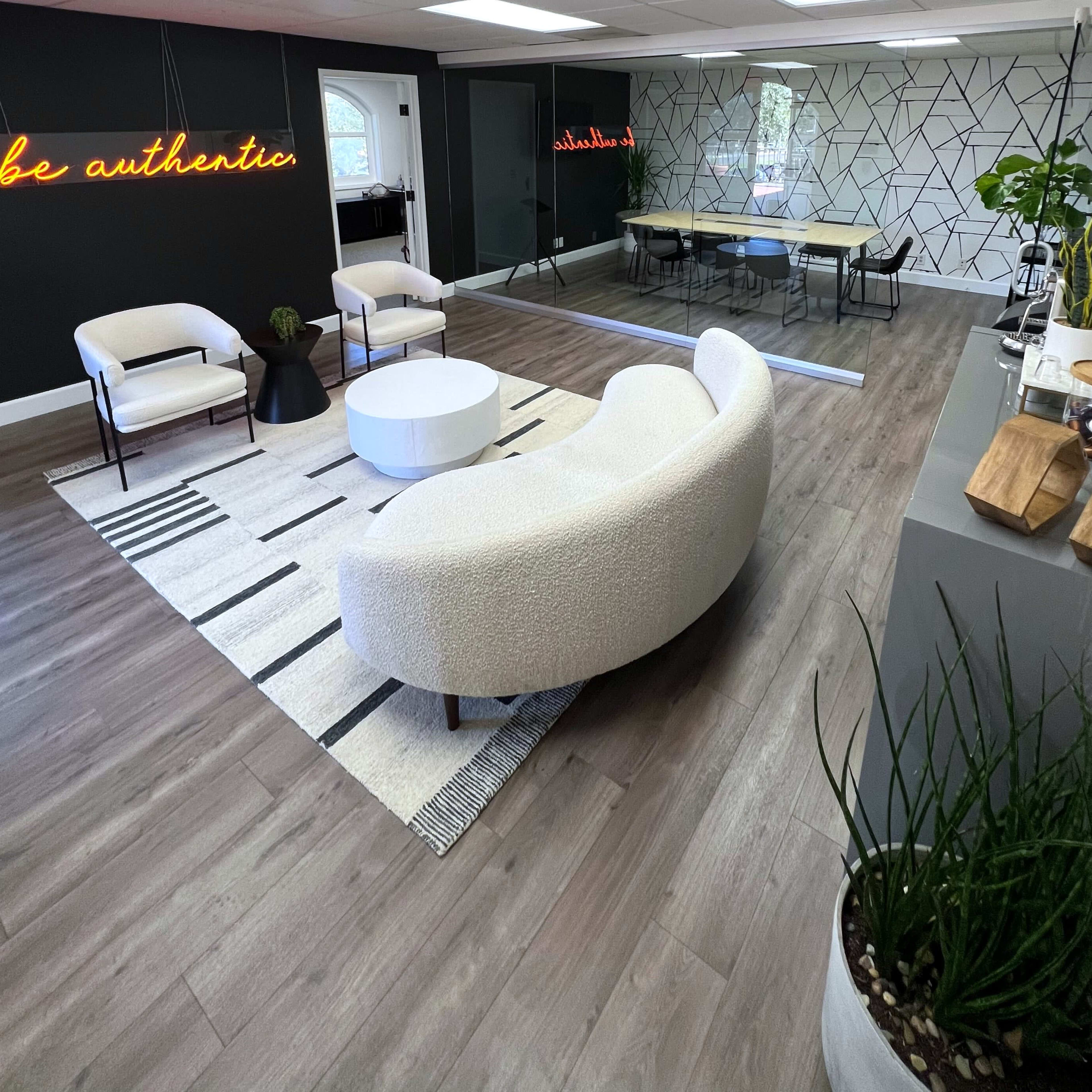A modern office space with a curved white sofa, two armchairs, a round coffee table on a patterned rug, and a large meeting table in a mirrored area.