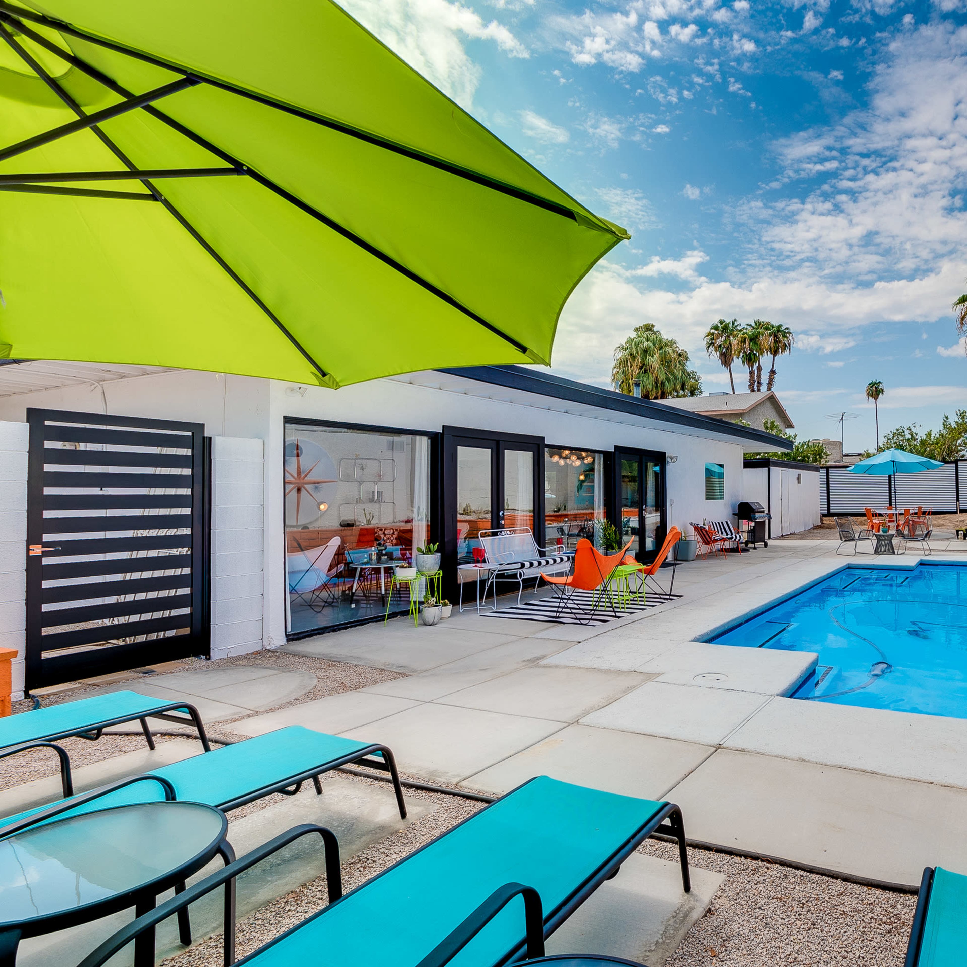 A modern backyard with a swimming pool, lounge chairs, and vibrant patio furniture surrounded by palm trees and a clear sky.