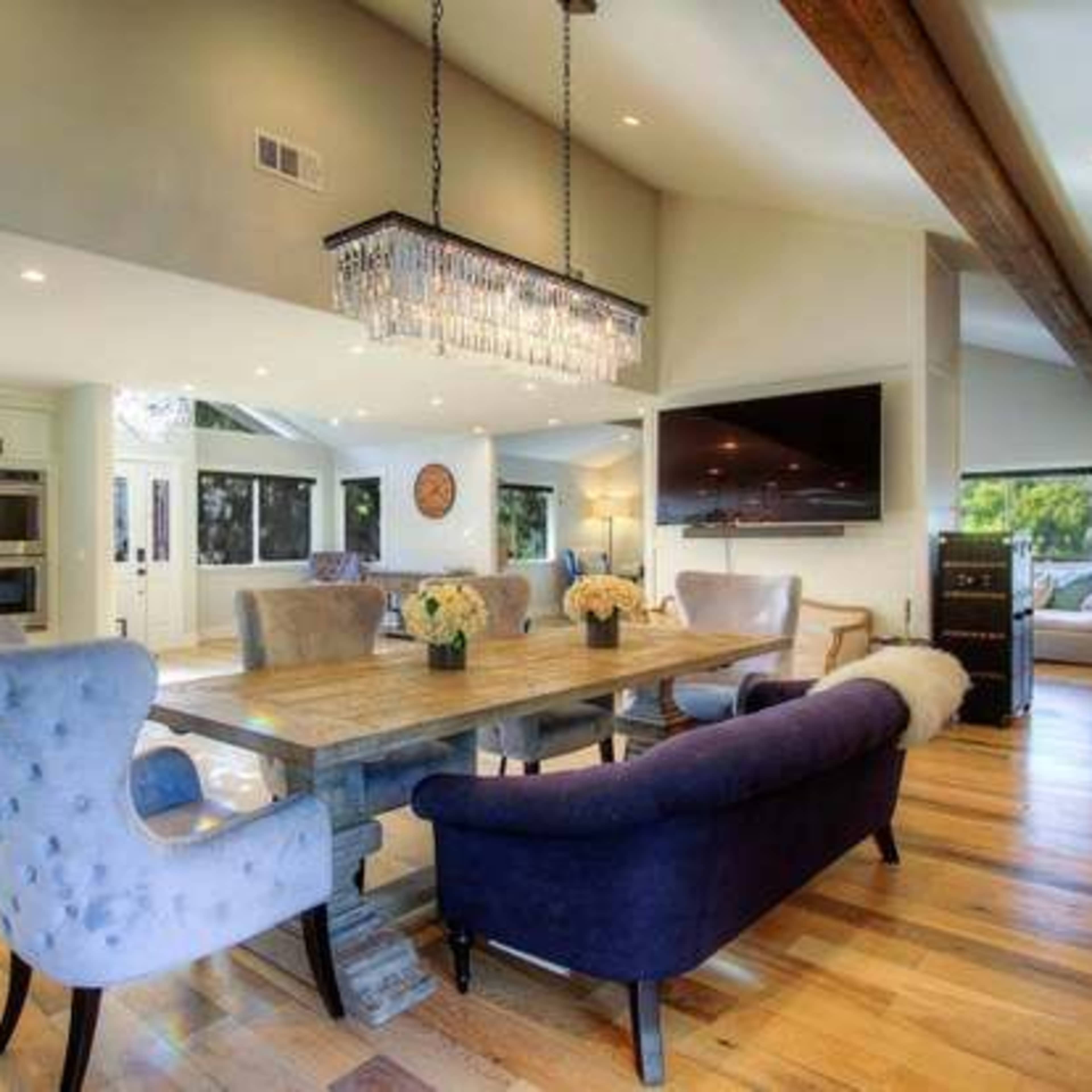 A modern dining area featuring a large wooden table surrounded by upholstered chairs, with a crystal chandelier overhead and a view of a bright kitchen in the background.