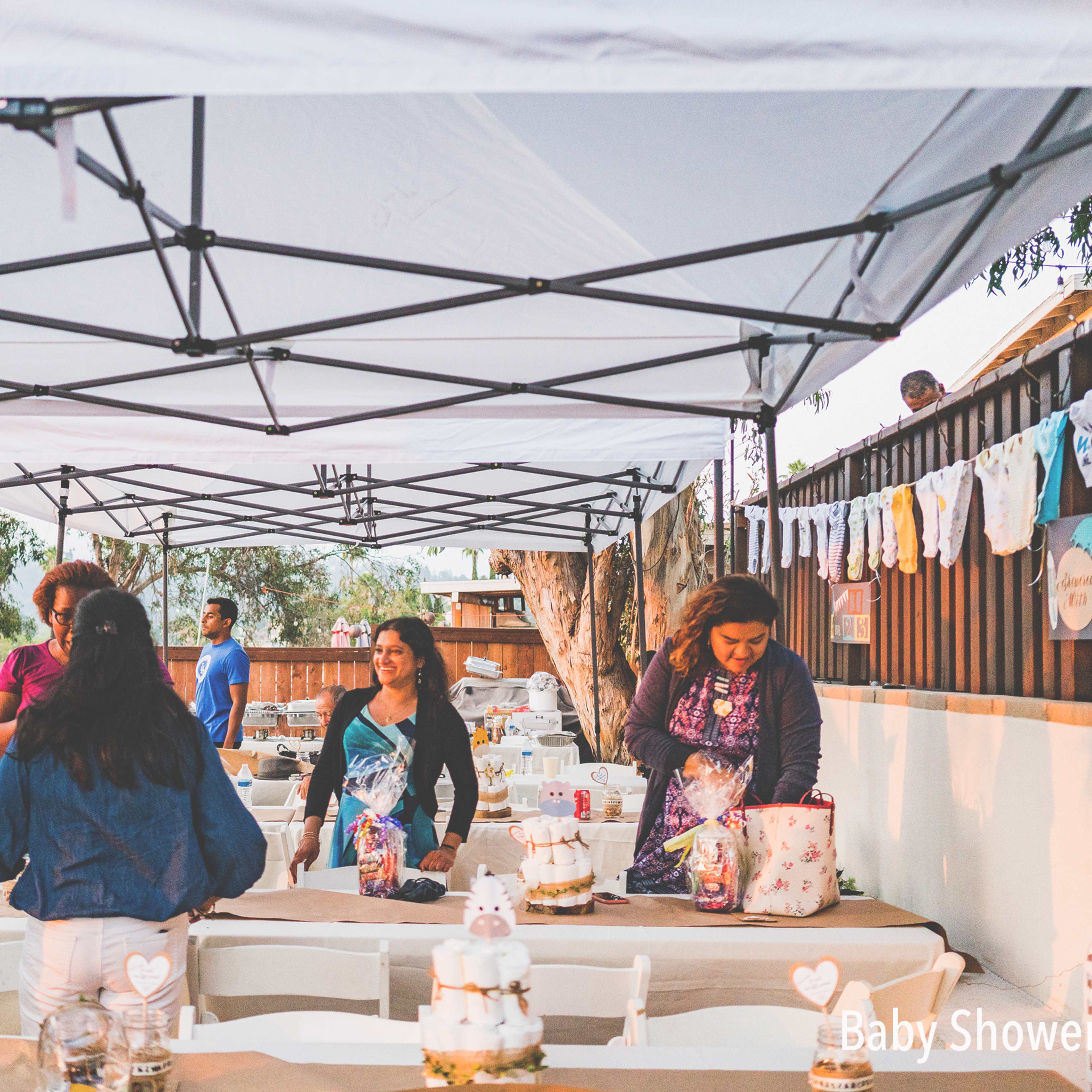 People are preparing a baby shower under tents with tables set up and decorations hanging on a fence.