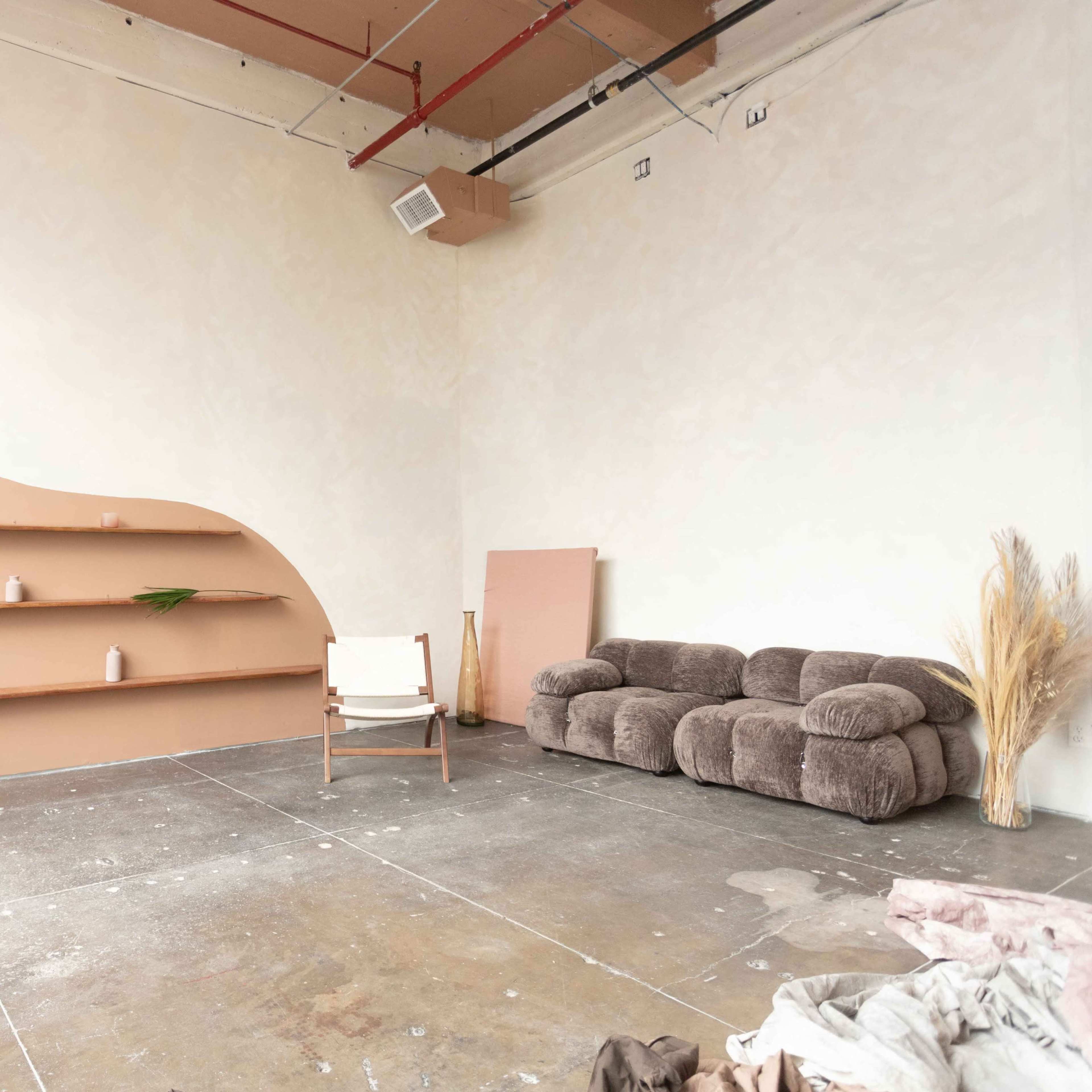 A minimalist interior space featuring a curved bookshelf, a cozy couch, a wooden chair, and unfinished flooring.