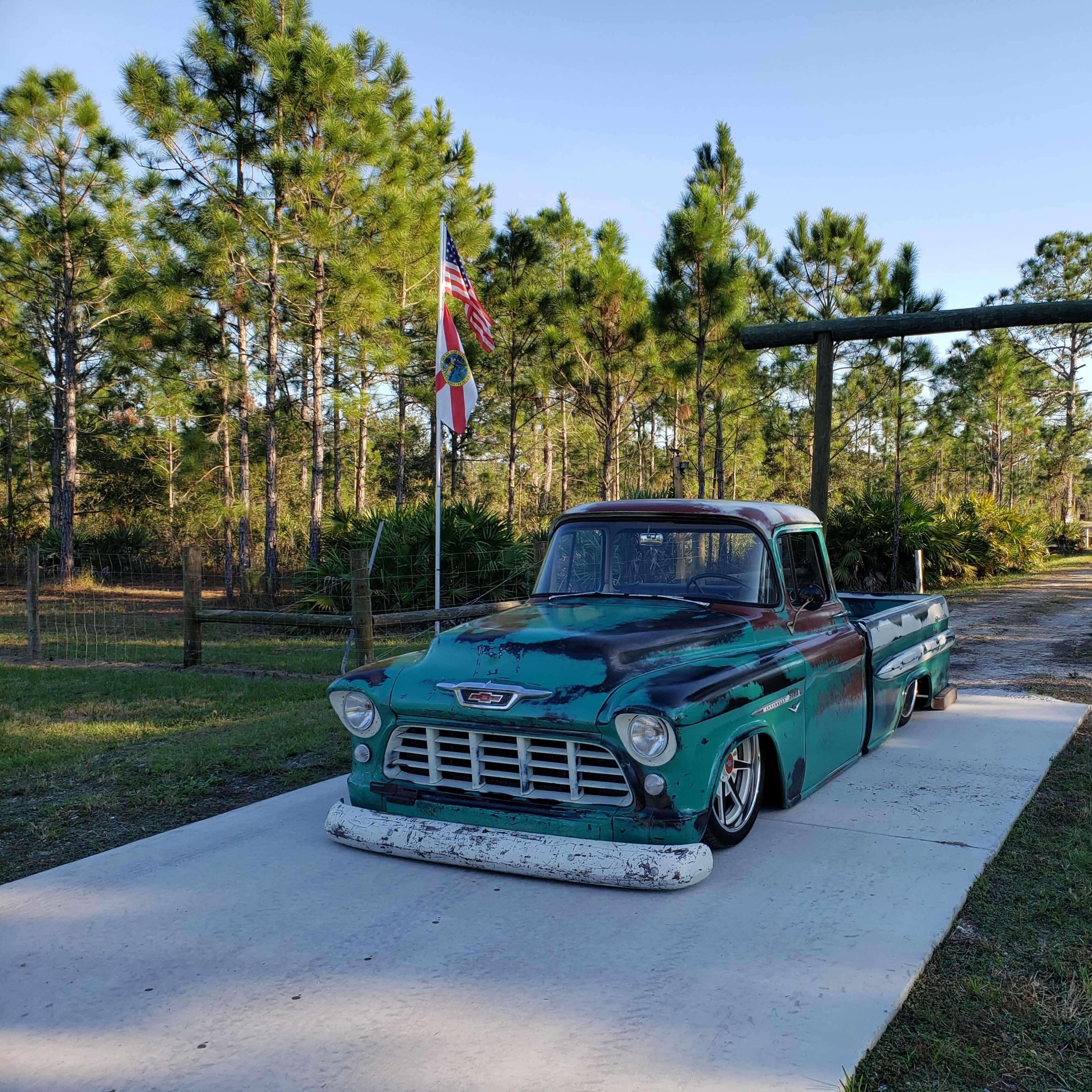 A vintage green pickup truck is parked on a concrete driveway leading to a tree-lined path with an American flag and a decorative signpost.