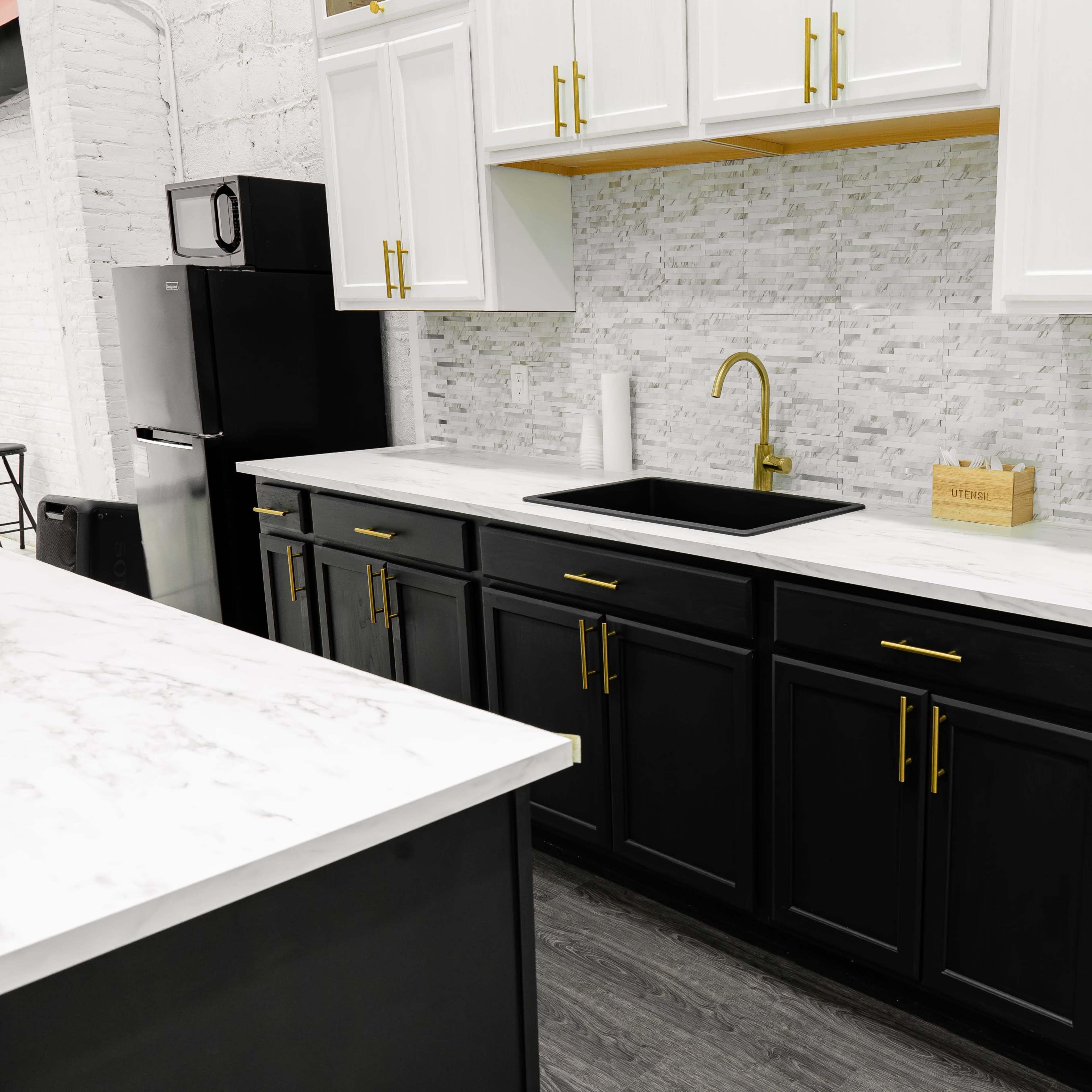 A modern kitchen with black lower cabinets, white upper cabinets, a marble countertop, and a gold faucet against a white and gray tiled backsplash.