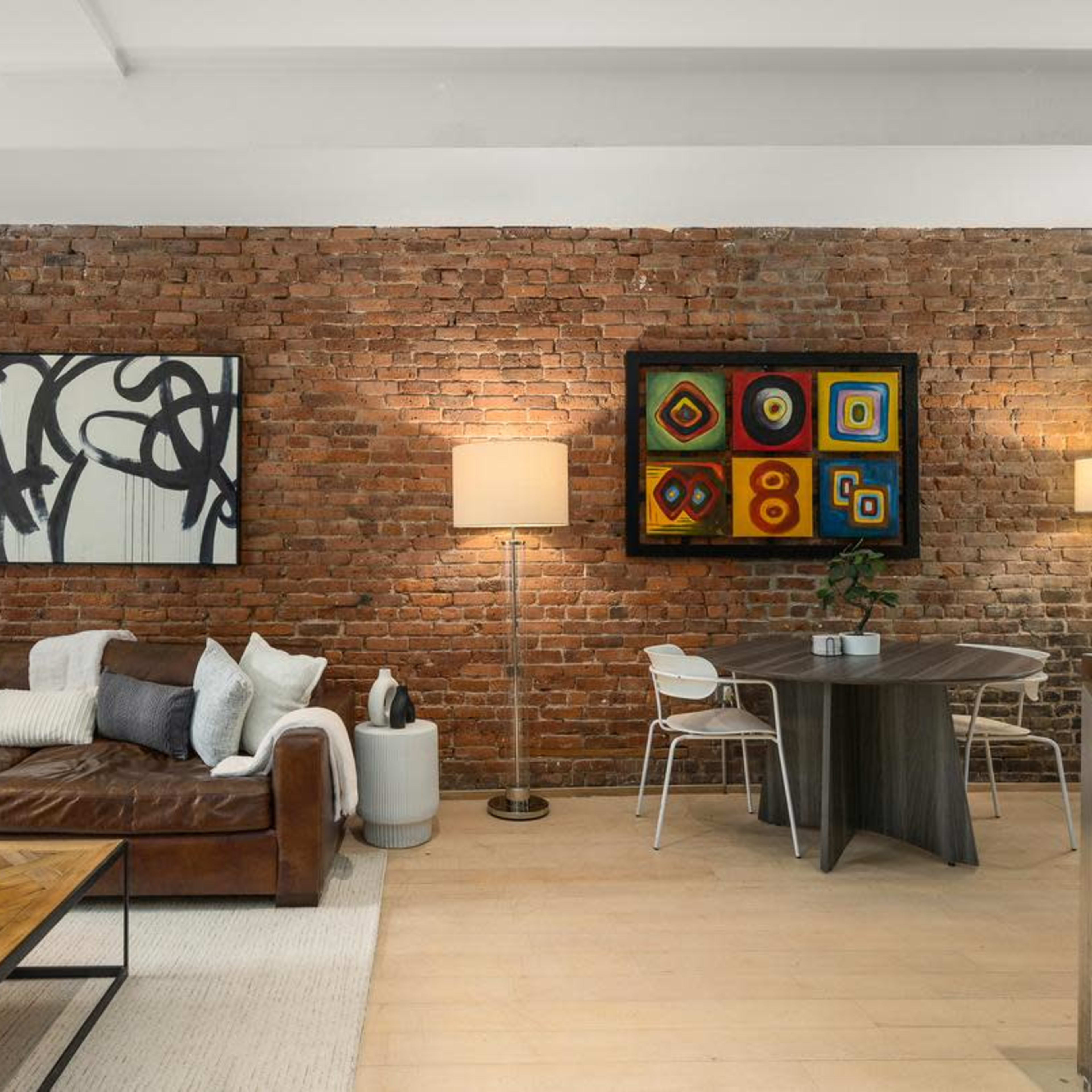 A living area with a brown leather sofa, two lamps, a wooden coffee table, and a dining table next to a brick wall adorned with colorful artwork.