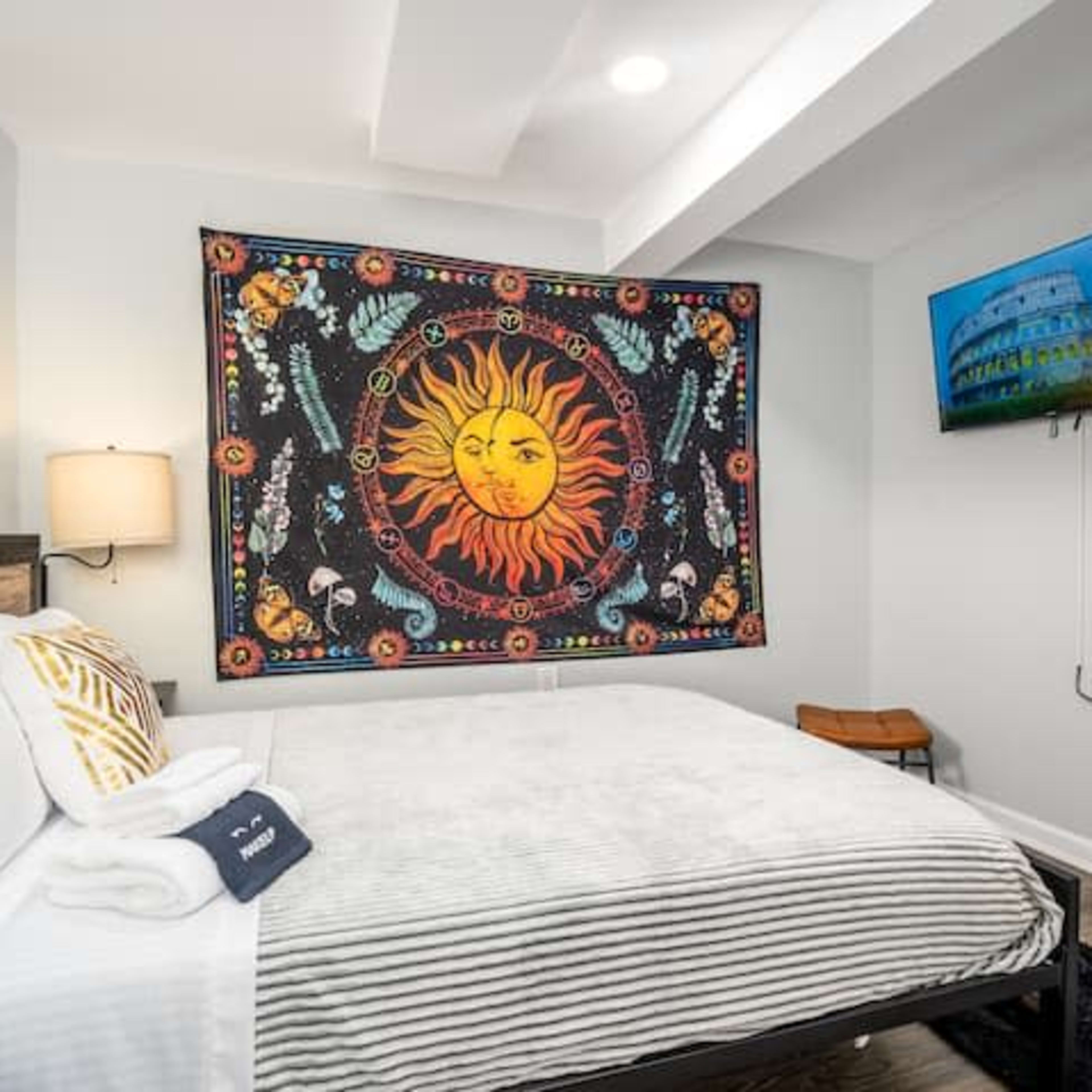 A bedroom with a bed, a tapestry featuring a sun design on the wall, and a television mounted nearby.