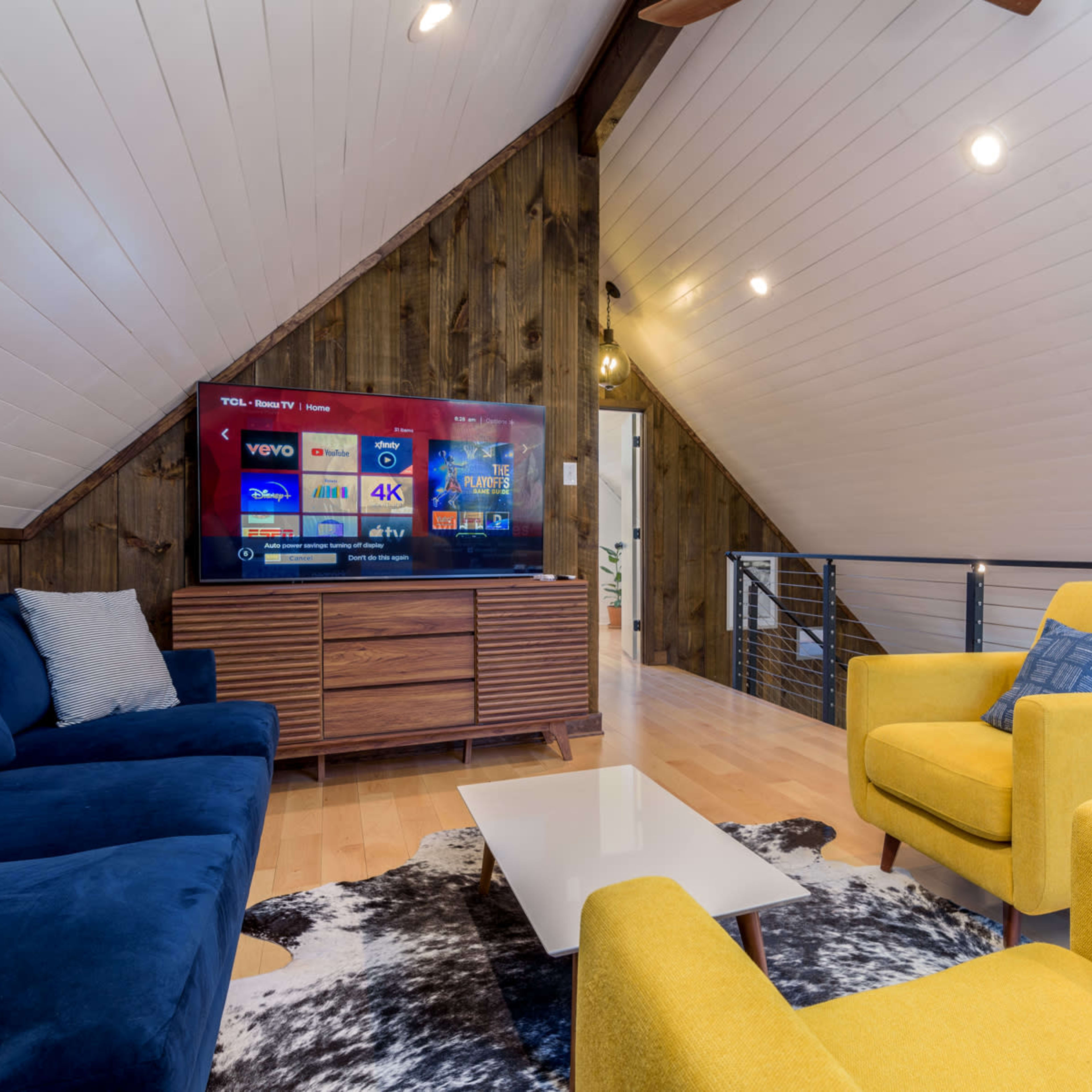 A cozy loft living area with a blue sectional sofa, yellow armchairs, and a wall-mounted television displaying various streaming options.
