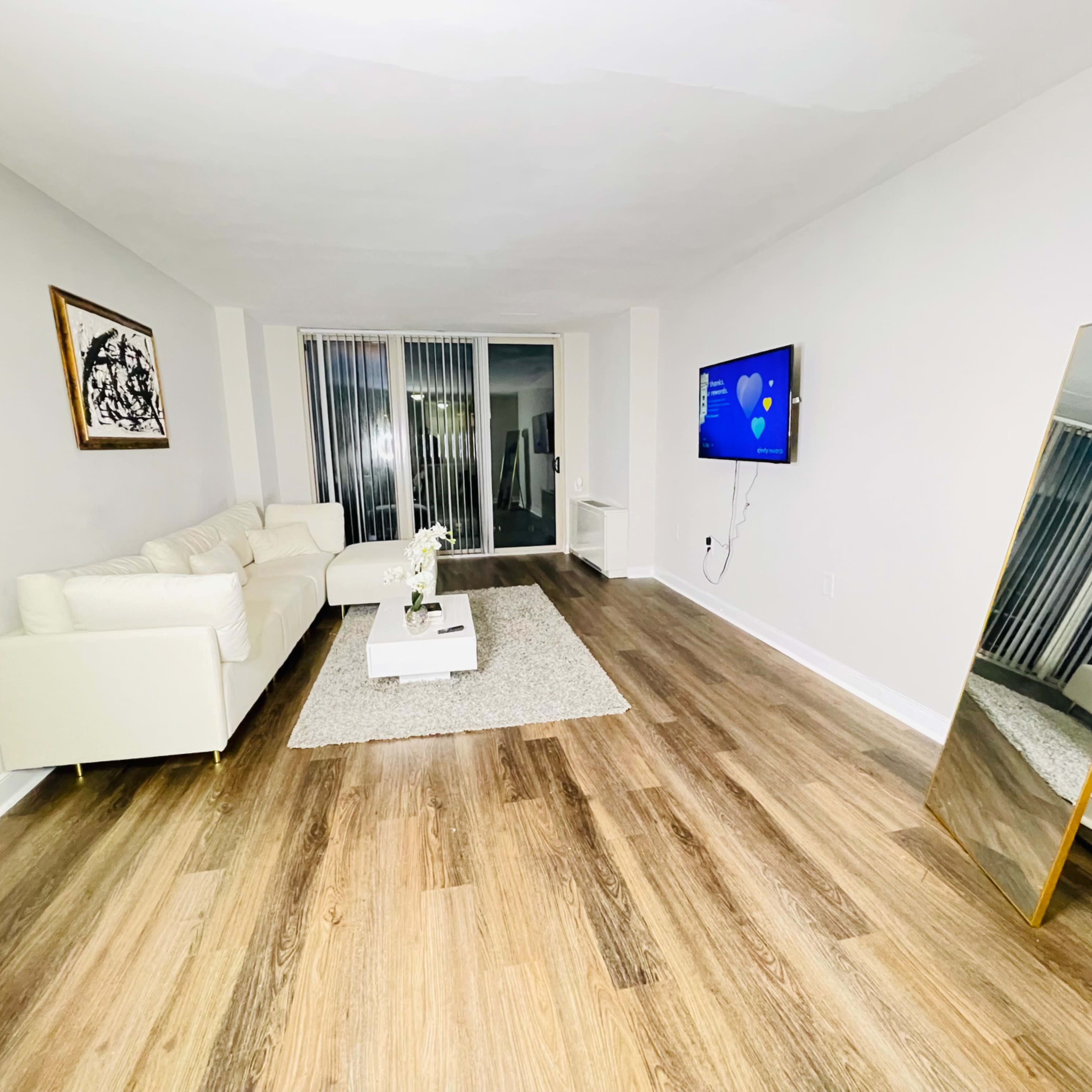 A modern, minimalist living room features a white sofa, a large mirror, a flat-screen TV, and hardwood flooring.