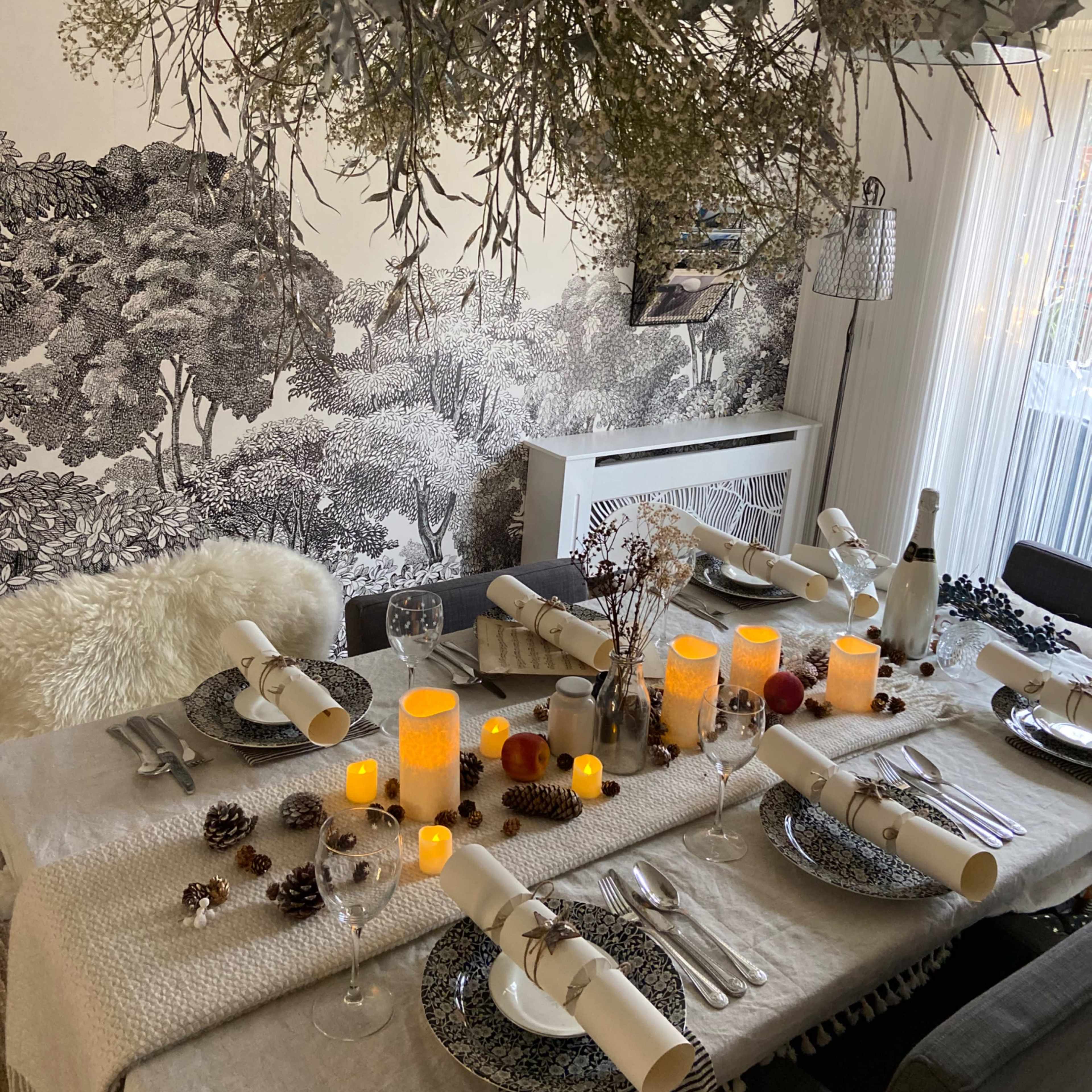 A decorated dining table is set with elegant tableware, candles, and pinecones, beneath a hanging arrangement of greenery, against a patterned wallpaper backdrop.