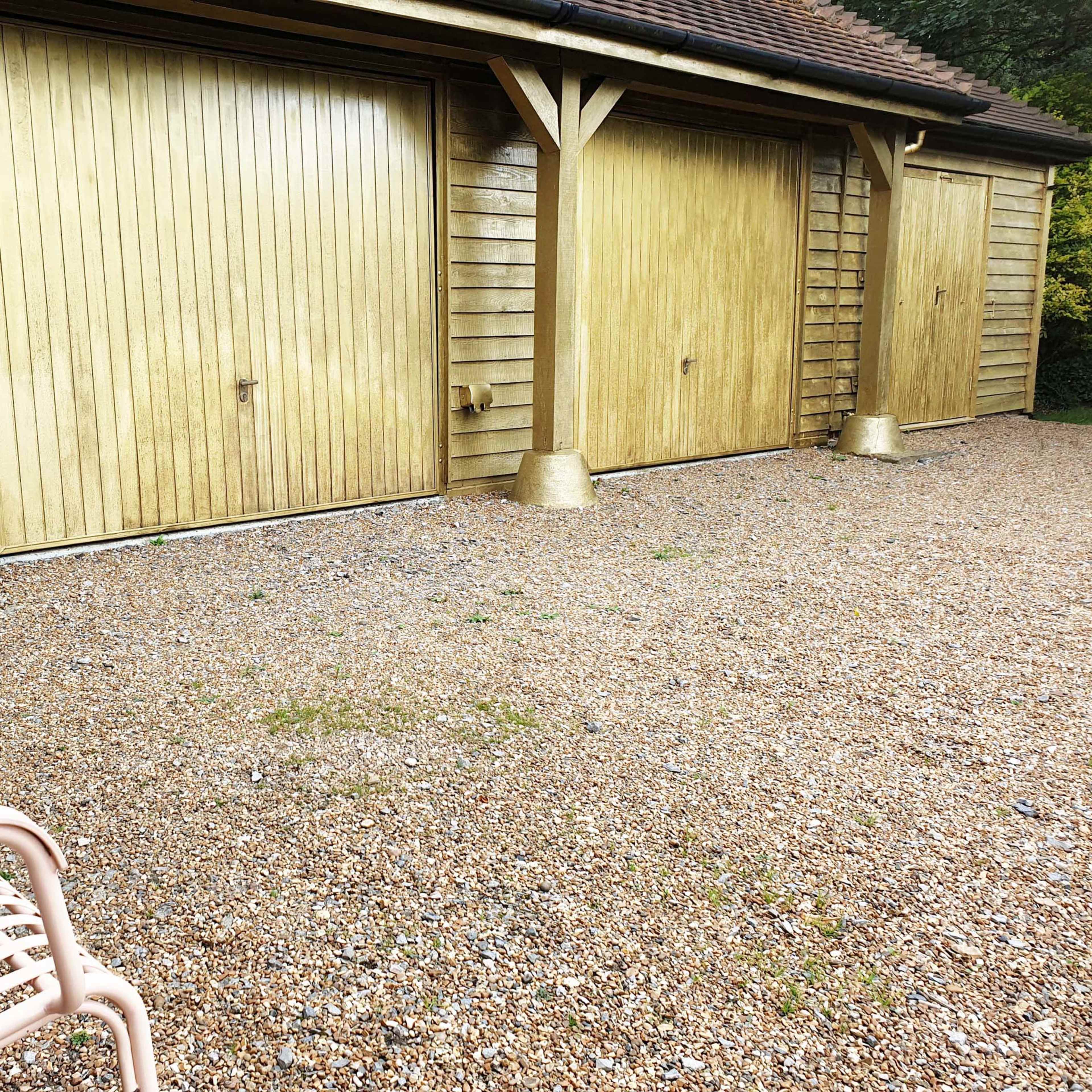 A gravel area in front of three wooden garage doors, with a metal bench positioned nearby.