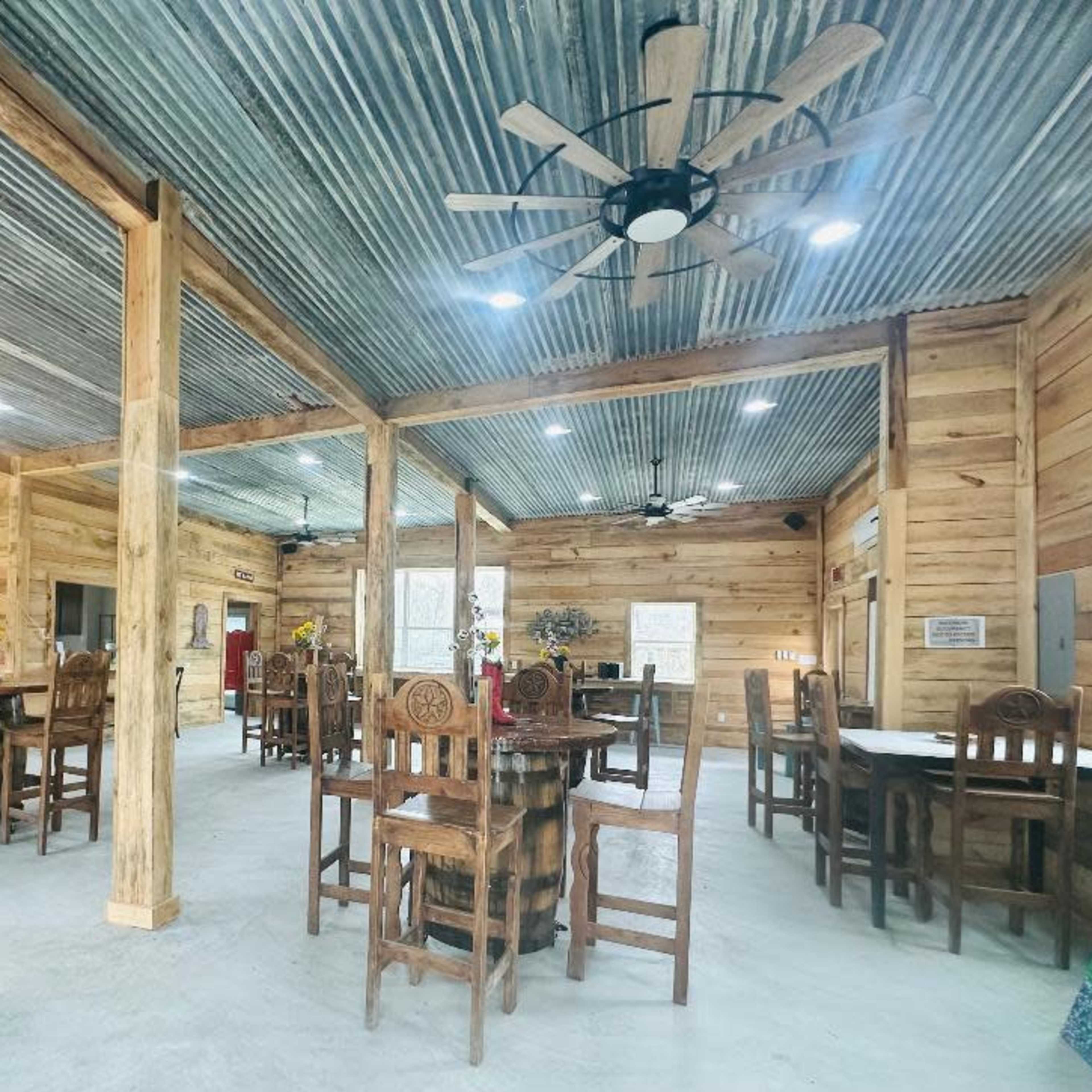 A spacious wooden interior with metal roofing, featuring several dining tables surrounded by chairs and a chandelier hanging from the ceiling.