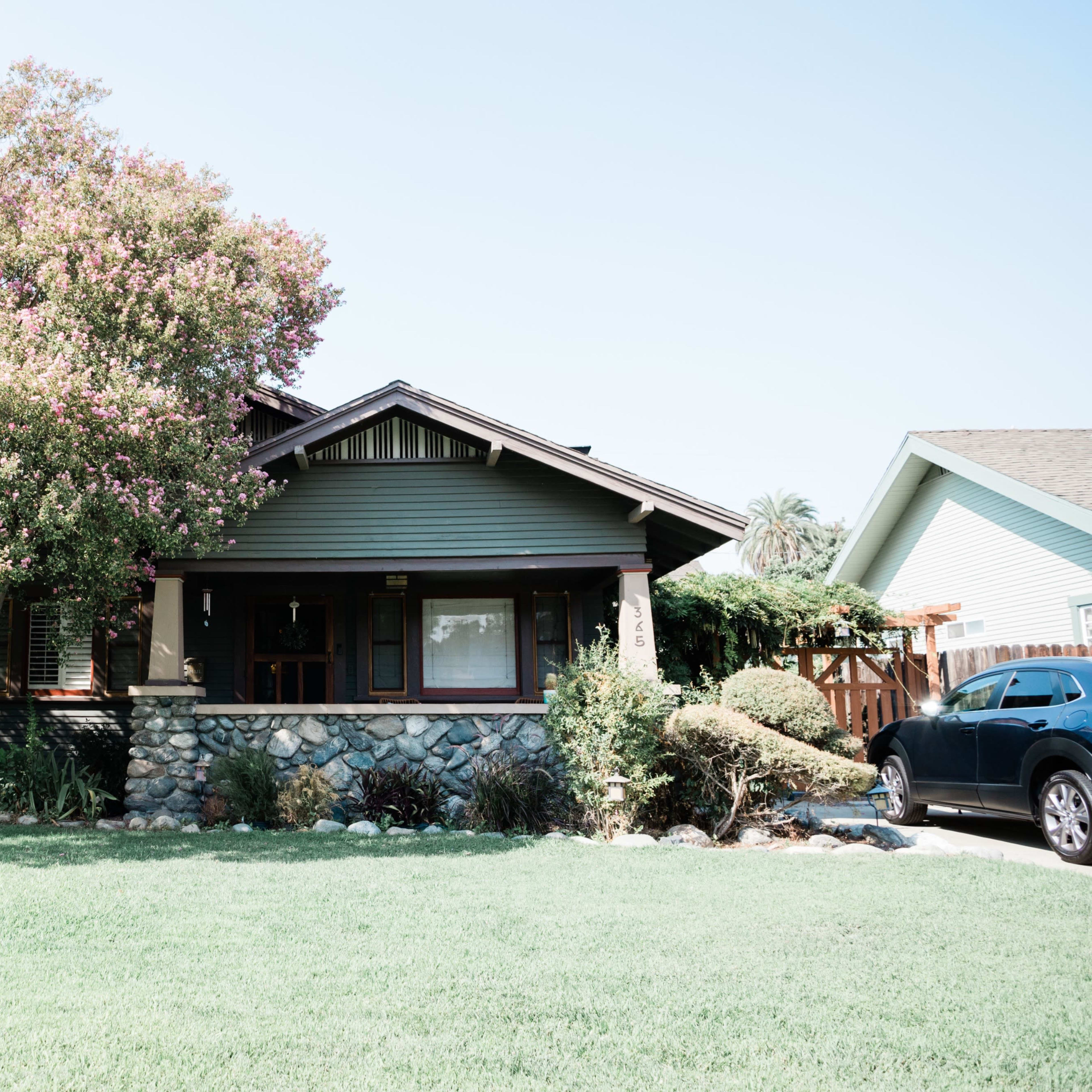 A craftsman-style house with a stone foundation is framed by a large flowering tree and a well-maintained lawn.