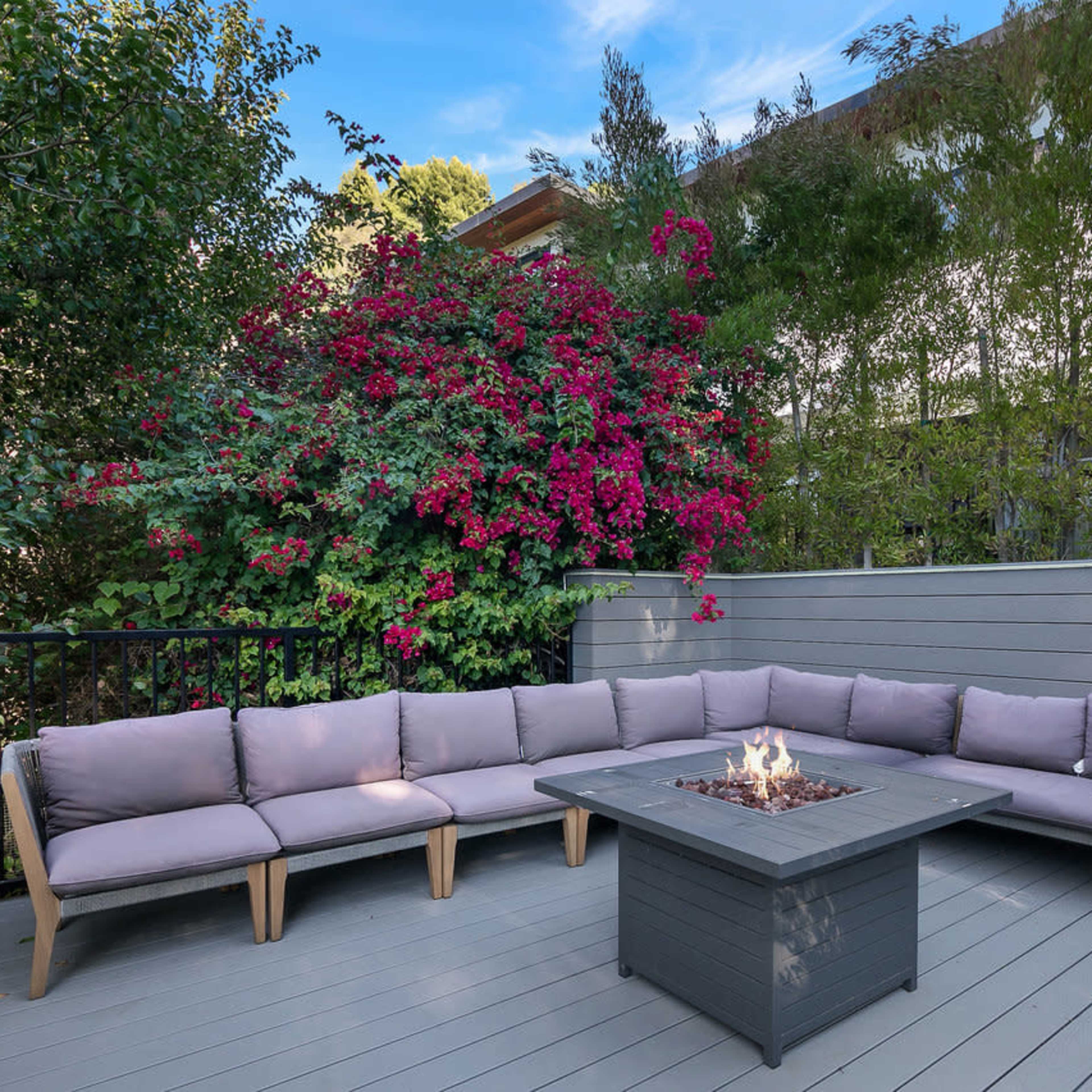 A recessed fire pit surrounded by a U-shaped outdoor sectional sofa is situated on a wooden deck, with vibrant flowering plants in the background.