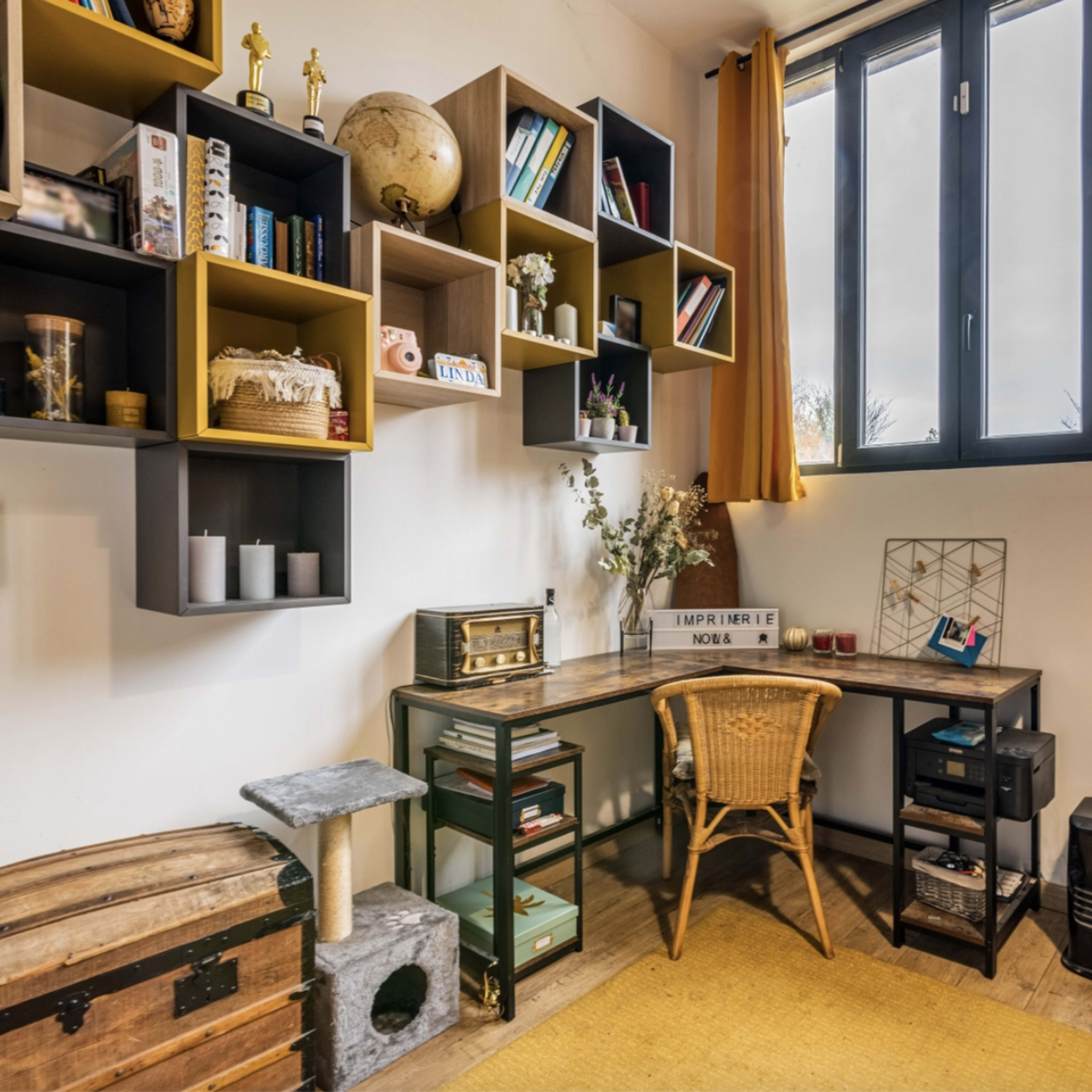 A cozy office space features a desk with a wicker chair, shelves filled with books and decor, and a large window with yellow curtains.