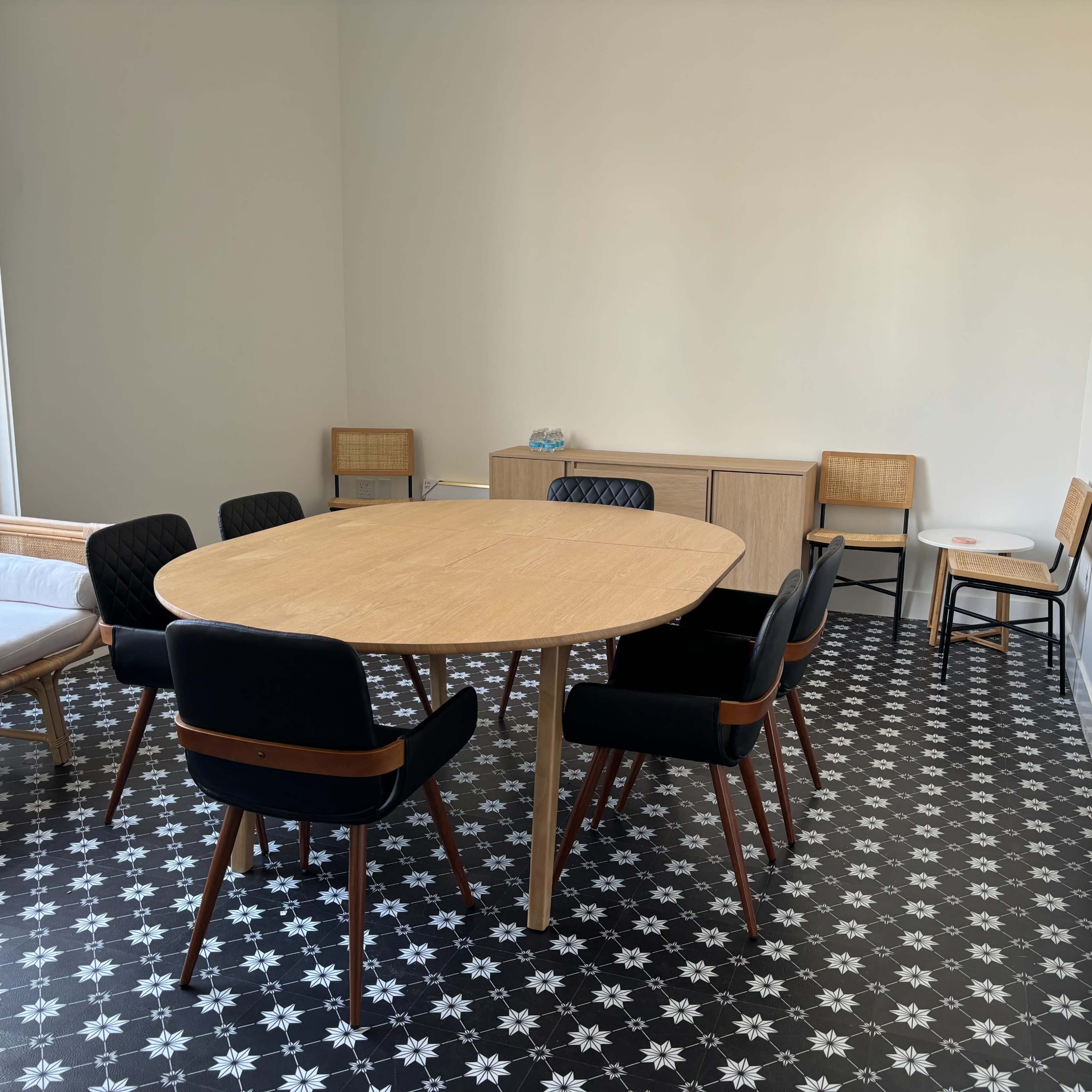 A round wooden table surrounded by black and wooden chairs is set in a minimally decorated room with patterned flooring.