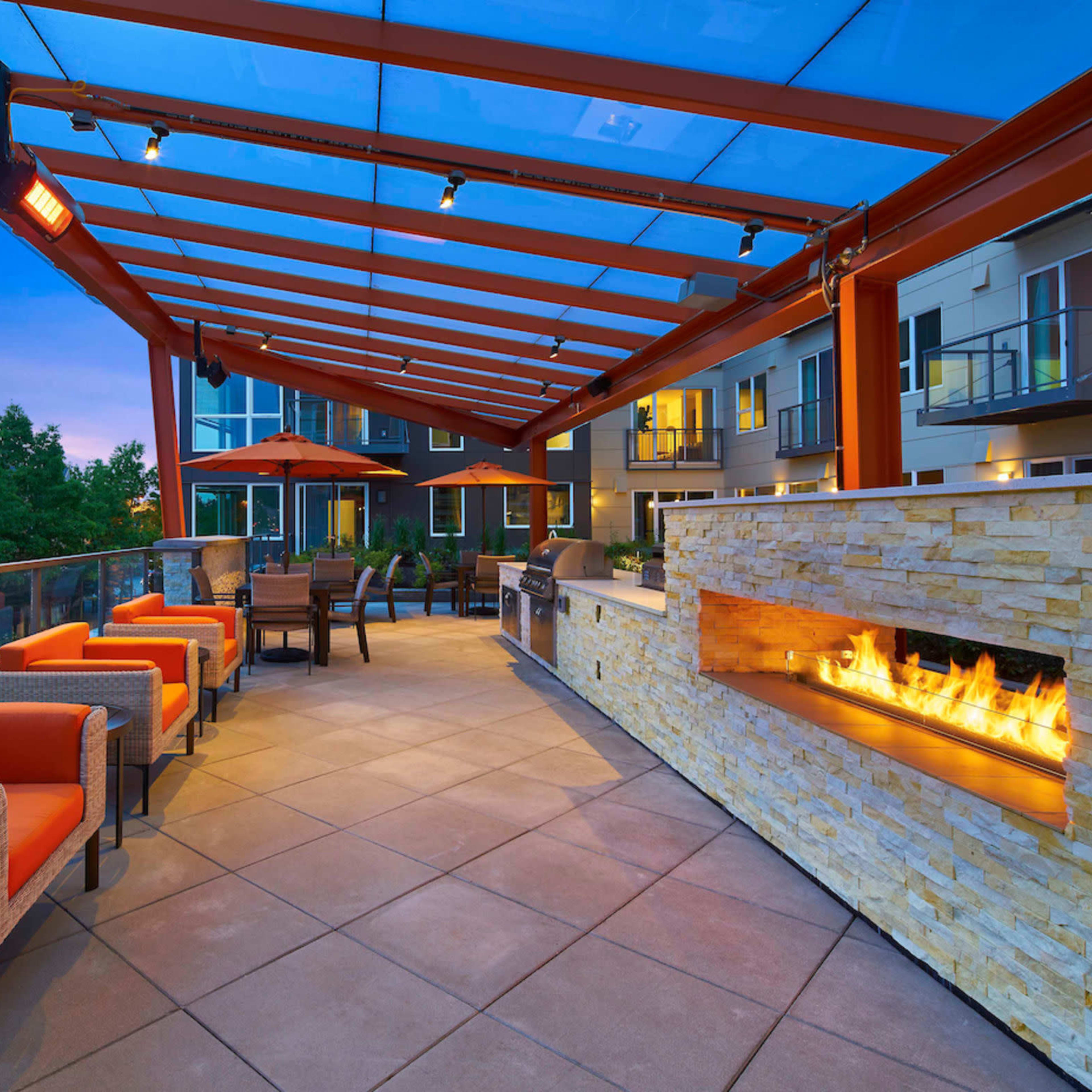 A modern outdoor seating area with orange chairs, a stone fireplace, and a grill under a red pergola.