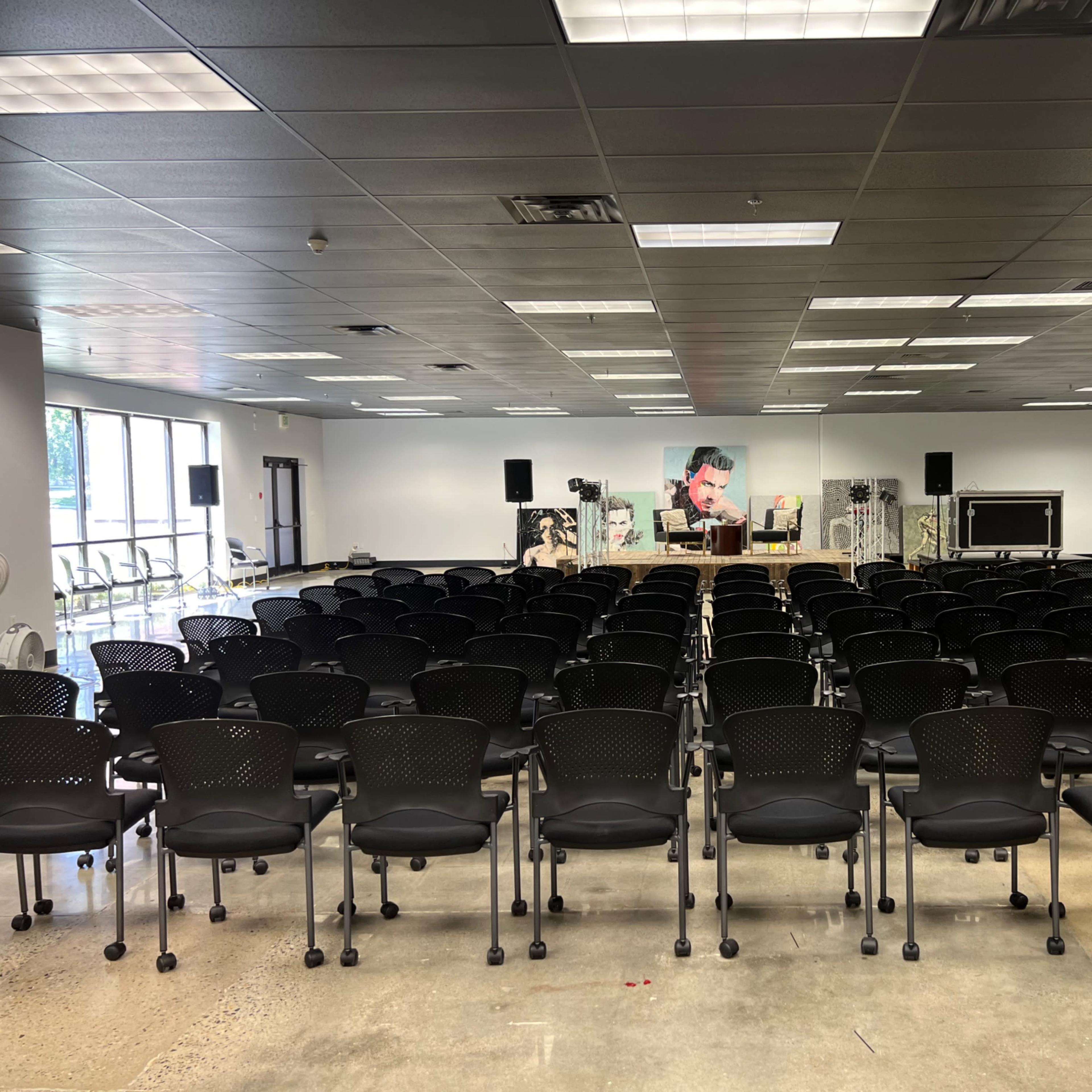A spacious event room is set up with rows of black chairs facing a stage adorned with a backdrop and two armchairs.