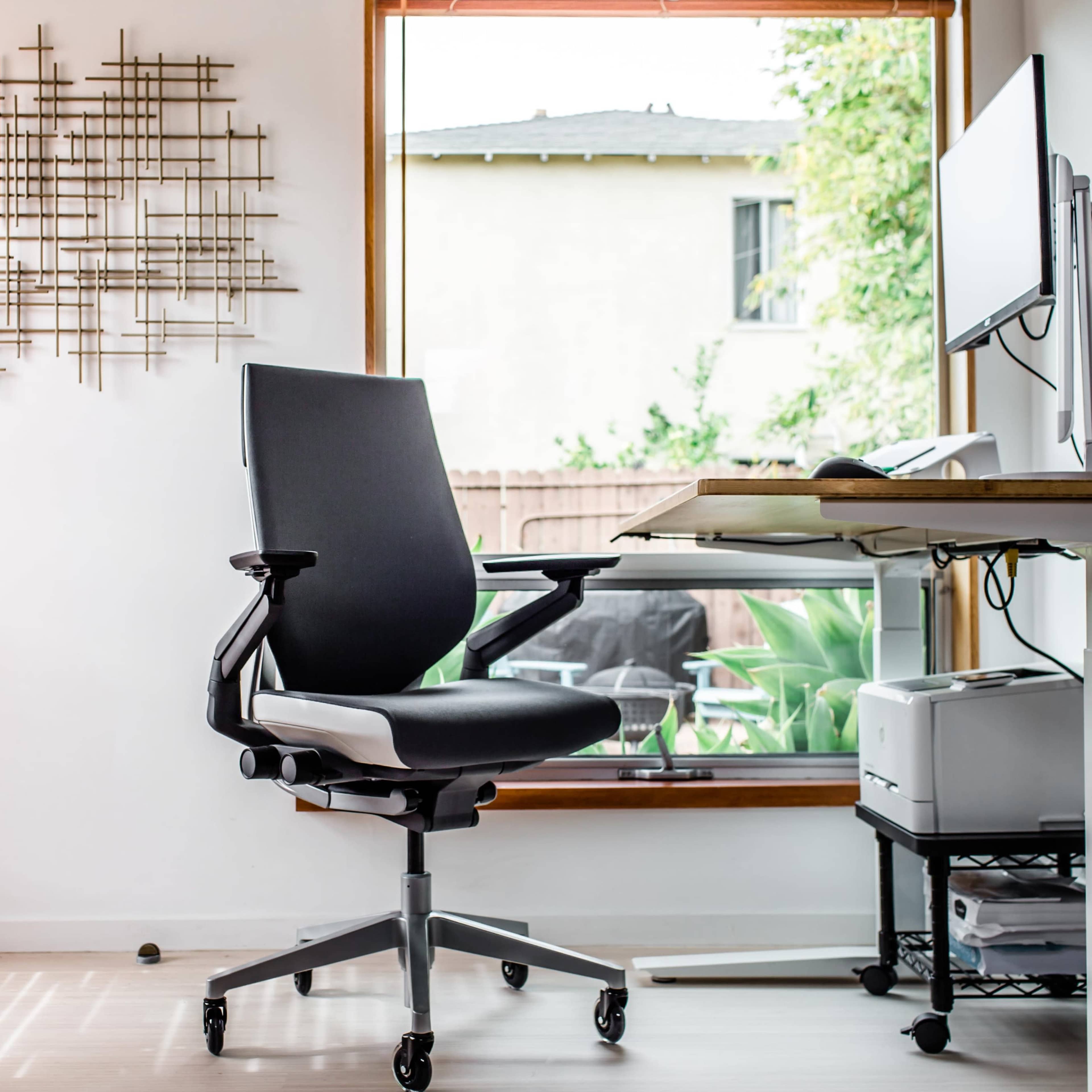 A black ergonomic chair is positioned in front of a desk with a computer monitor, beside a large window with wooden blinds.
