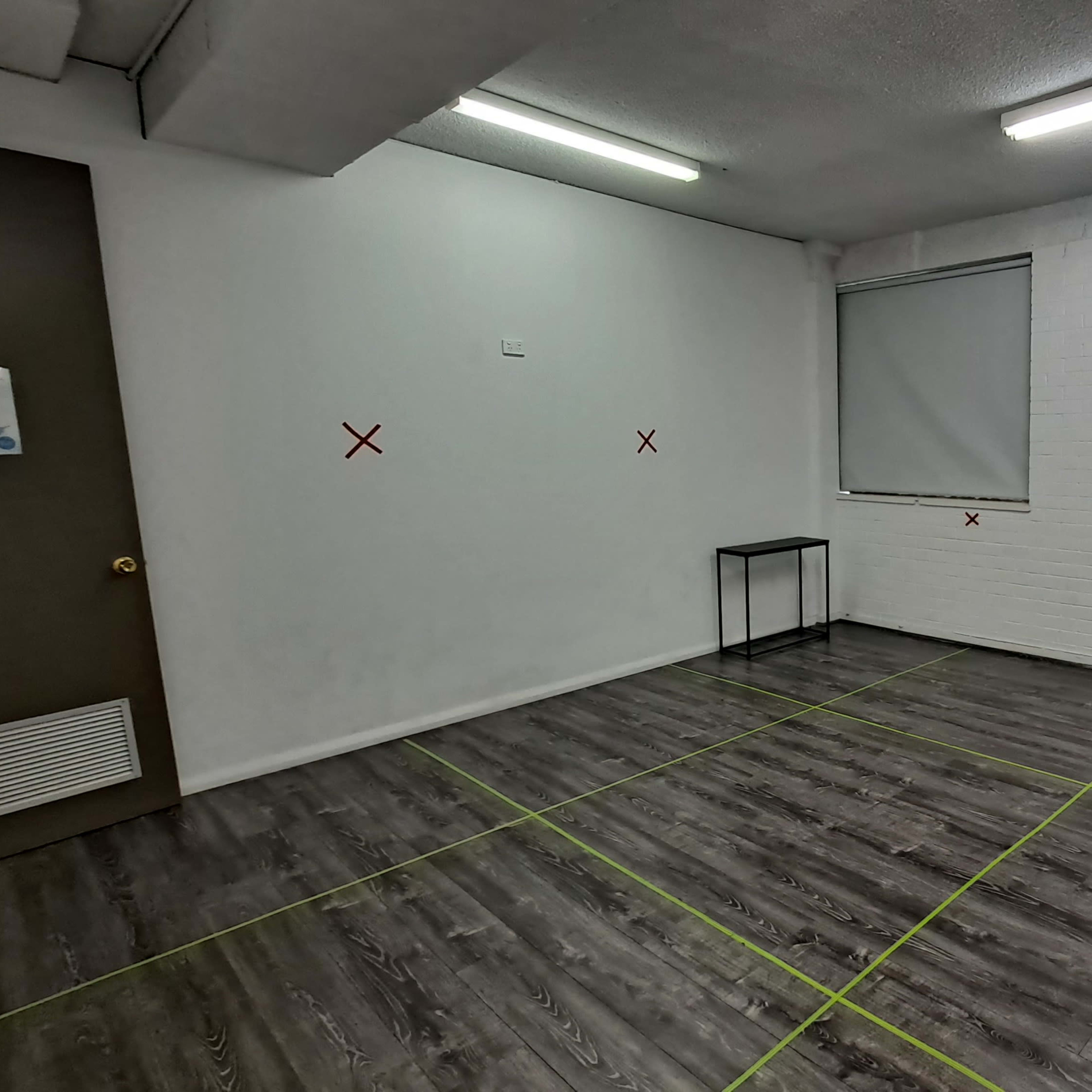 An empty room with white walls, a window covered by a blind, a small black table, and green tape marking boundaries on the floor.