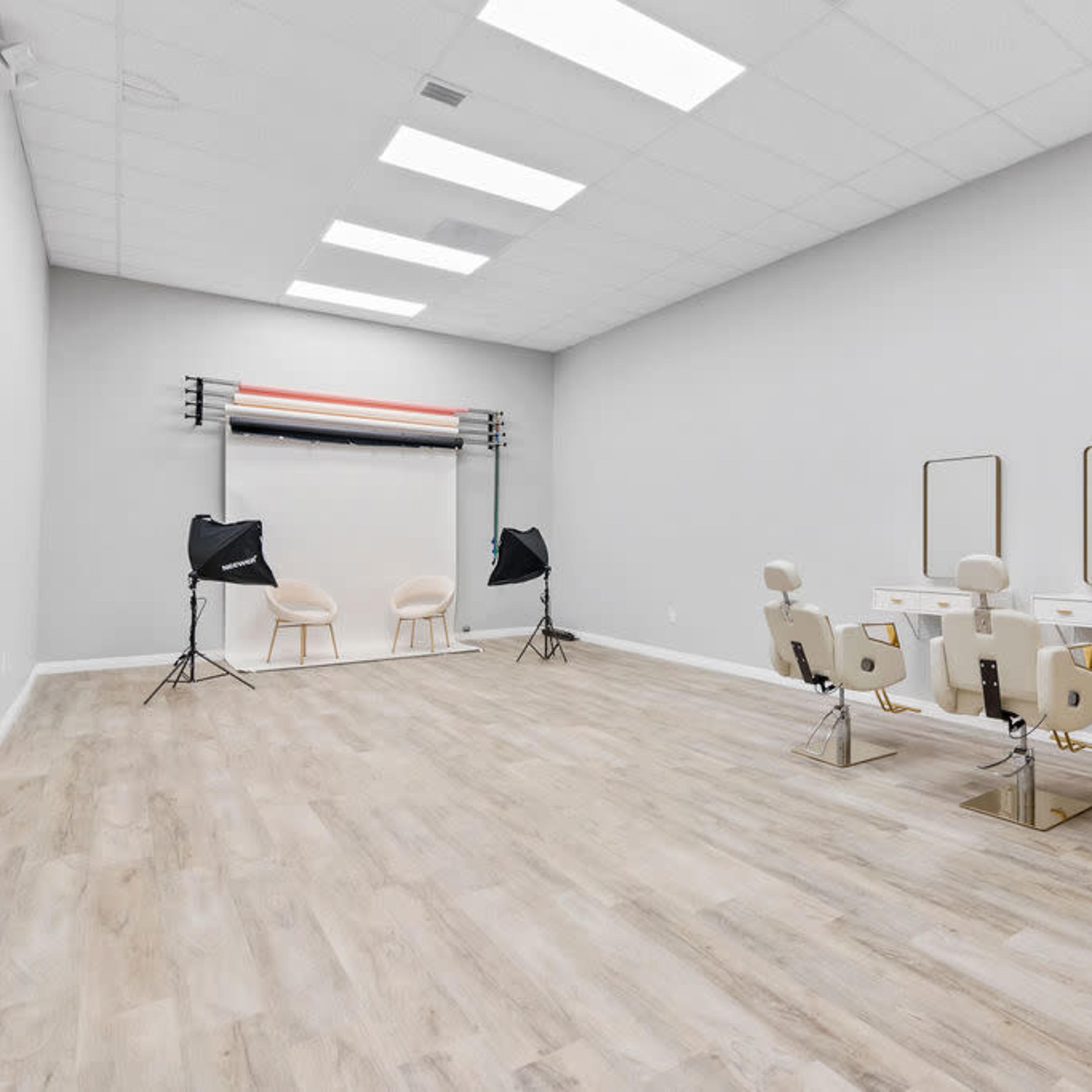 A spacious, minimalist studio interior with two hair styling stations, a backdrop setup, and two photography lights.
