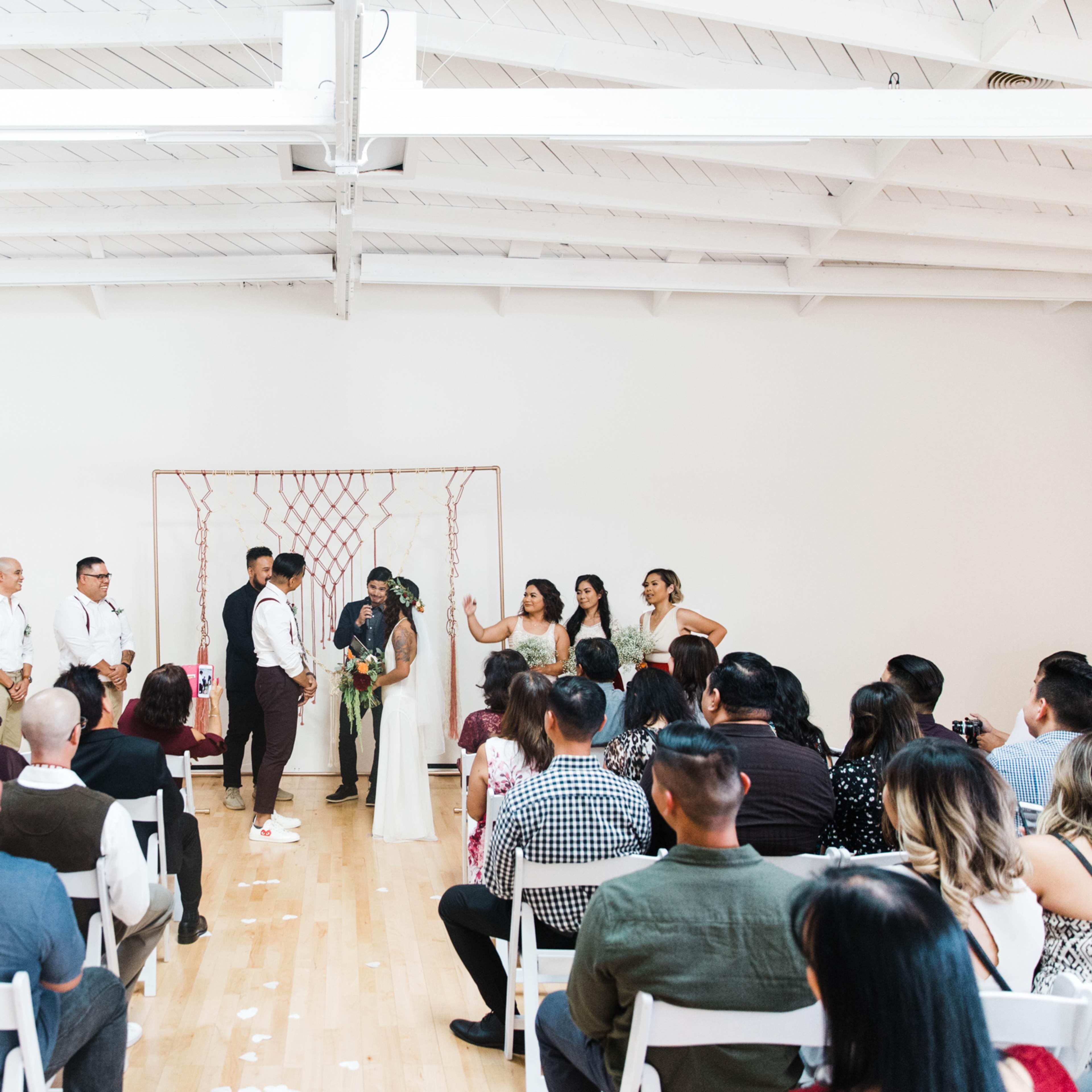 A wedding ceremony takes place indoors with a couple standing at the altar surrounded by their wedding party and a seated audience.