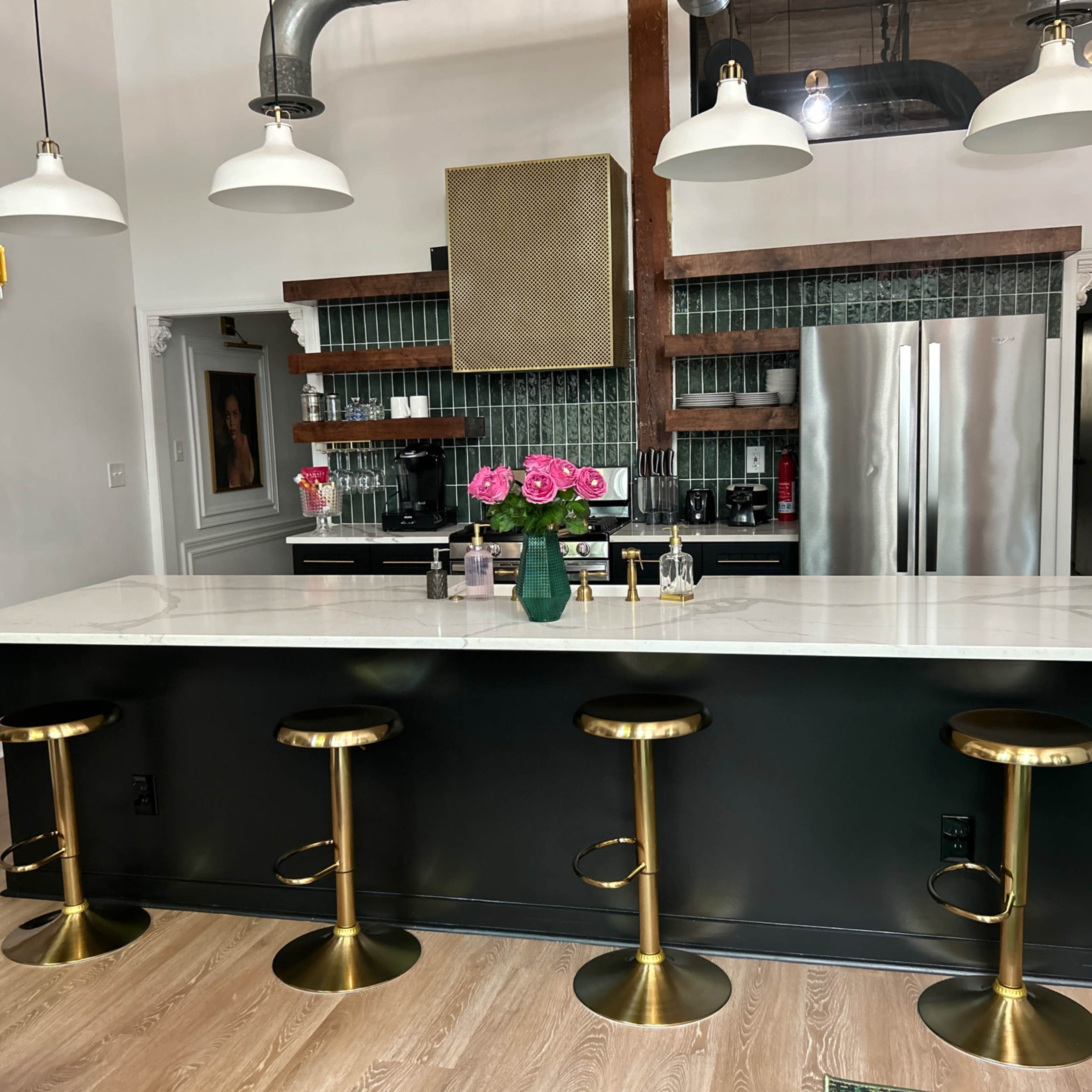 A modern kitchen features a marble countertop island with four gold bar stools, a green tiled backsplash, and stainless steel appliances.