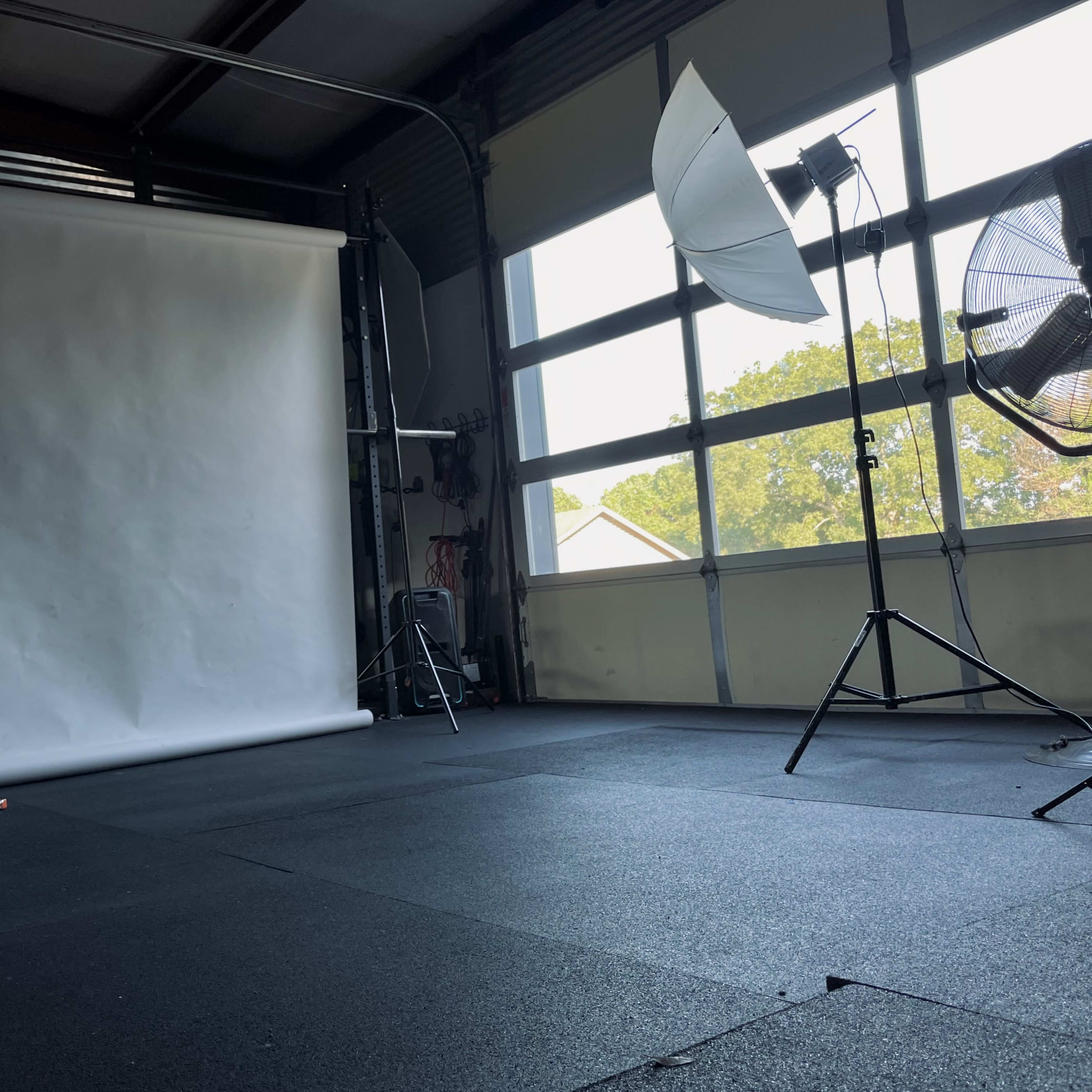 A photography studio with two softbox lights, a backdrop stand with a white paper roll, and two large fans positioned near a window.