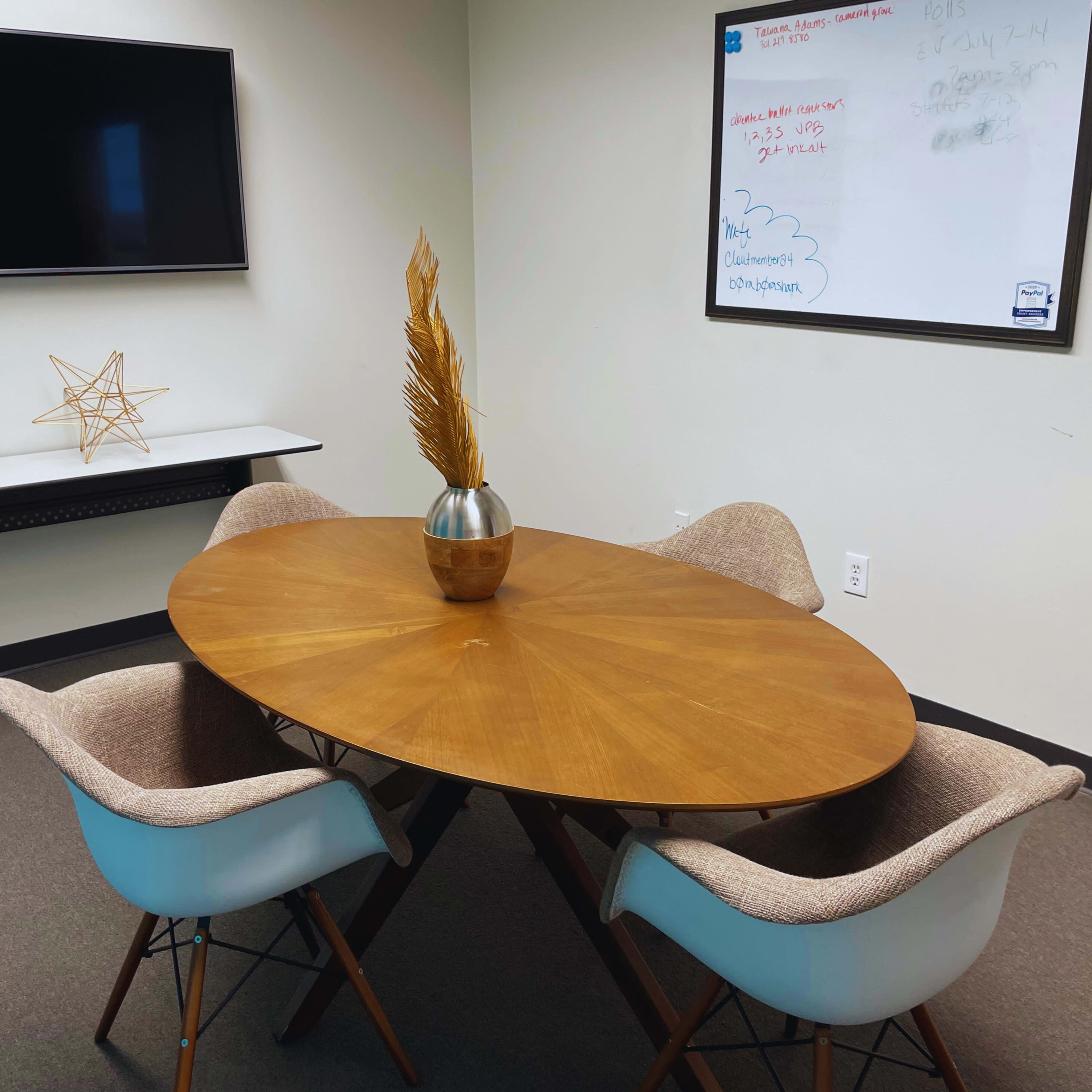 A modern conference room features a wooden oval table with a decorative vase and dried plants, surrounded by upholstered chairs and equipped with a wall-mounted TV and whiteboard.