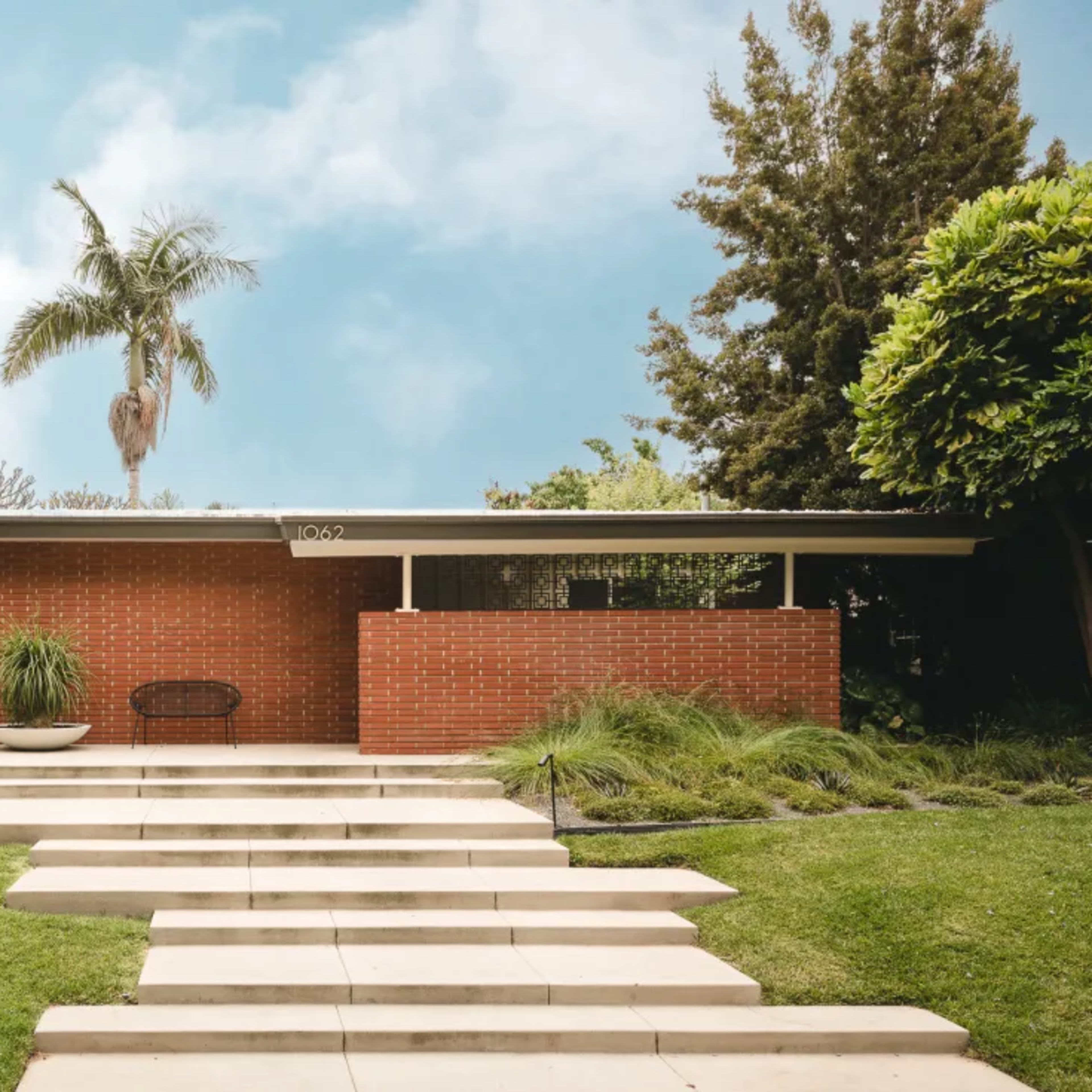 A modern brick house with a flat roof is set in a landscaped yard featuring a pathway and ornamental plants.