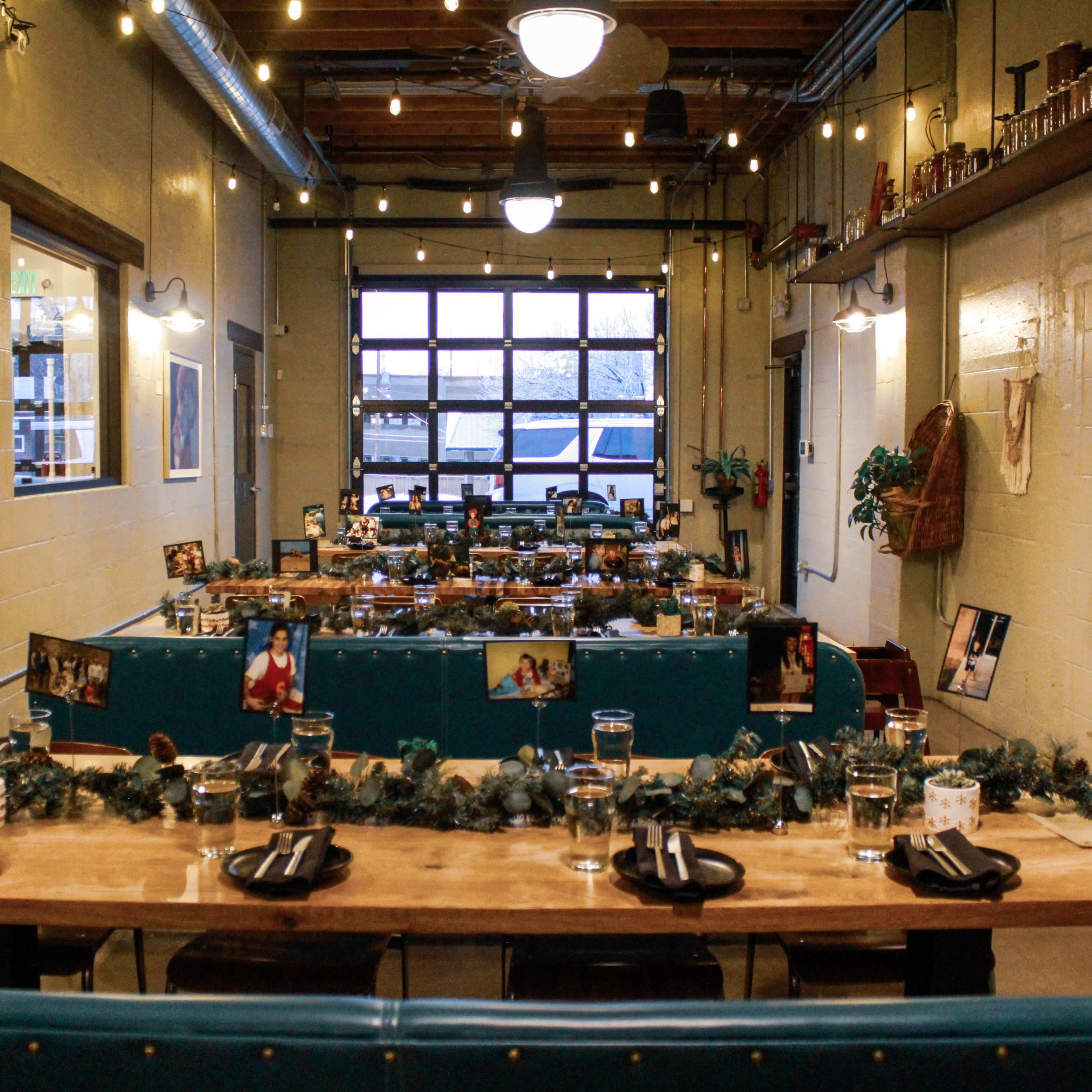 A decorated dining space with wooden tables set for a meal, string lights overhead, and a large garage door at the end of the room.
