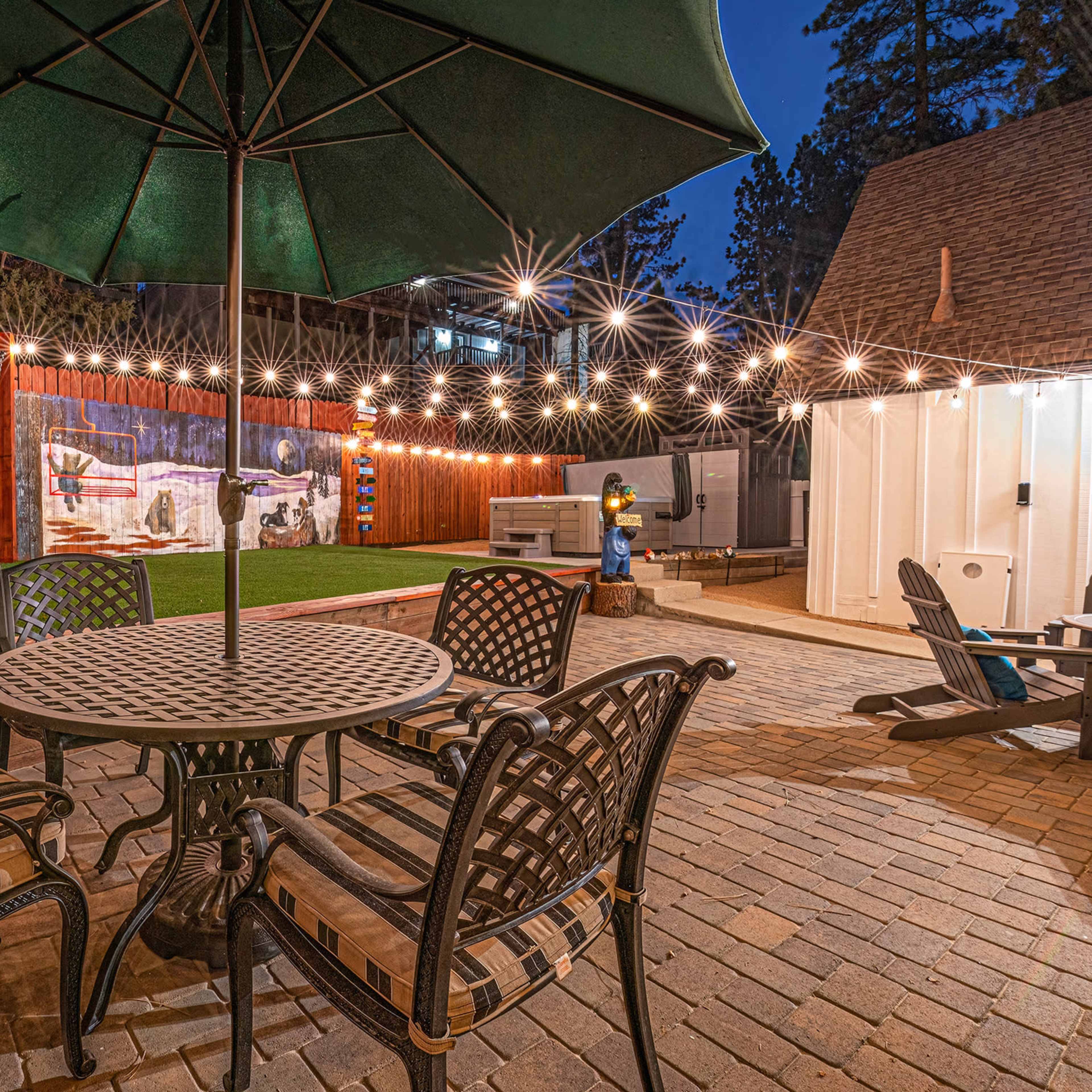 A patio scene features a round table with chairs, an umbrella, a hammock against a wooden fence, and string lights illuminating a grassy area.