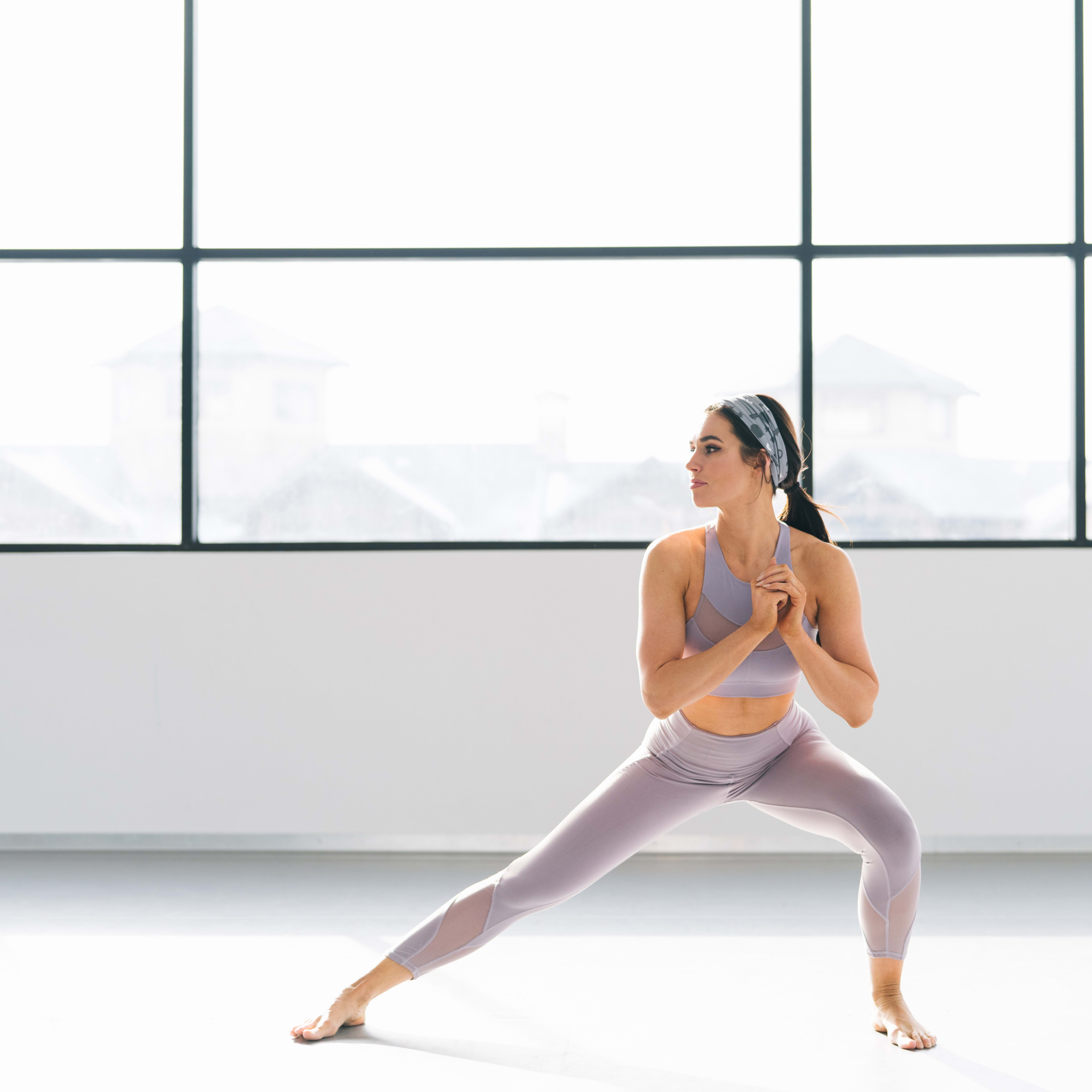 A woman in athletic wear performs a lunging exercise in a brightly lit studio with large windows.