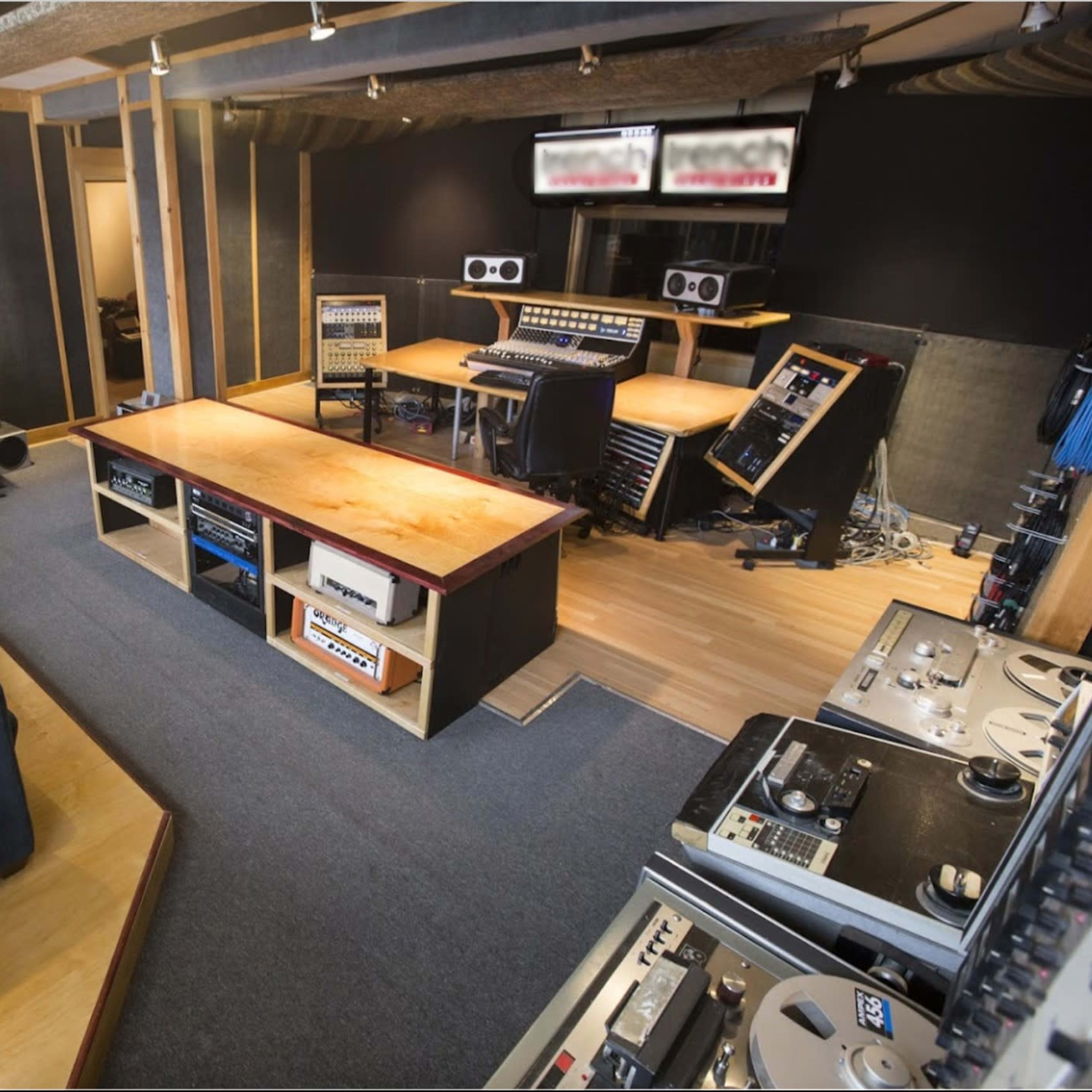 A recording studio featuring a central wooden table surrounded by audio equipment, mixing consoles, and a blue couch in a well-lit space.