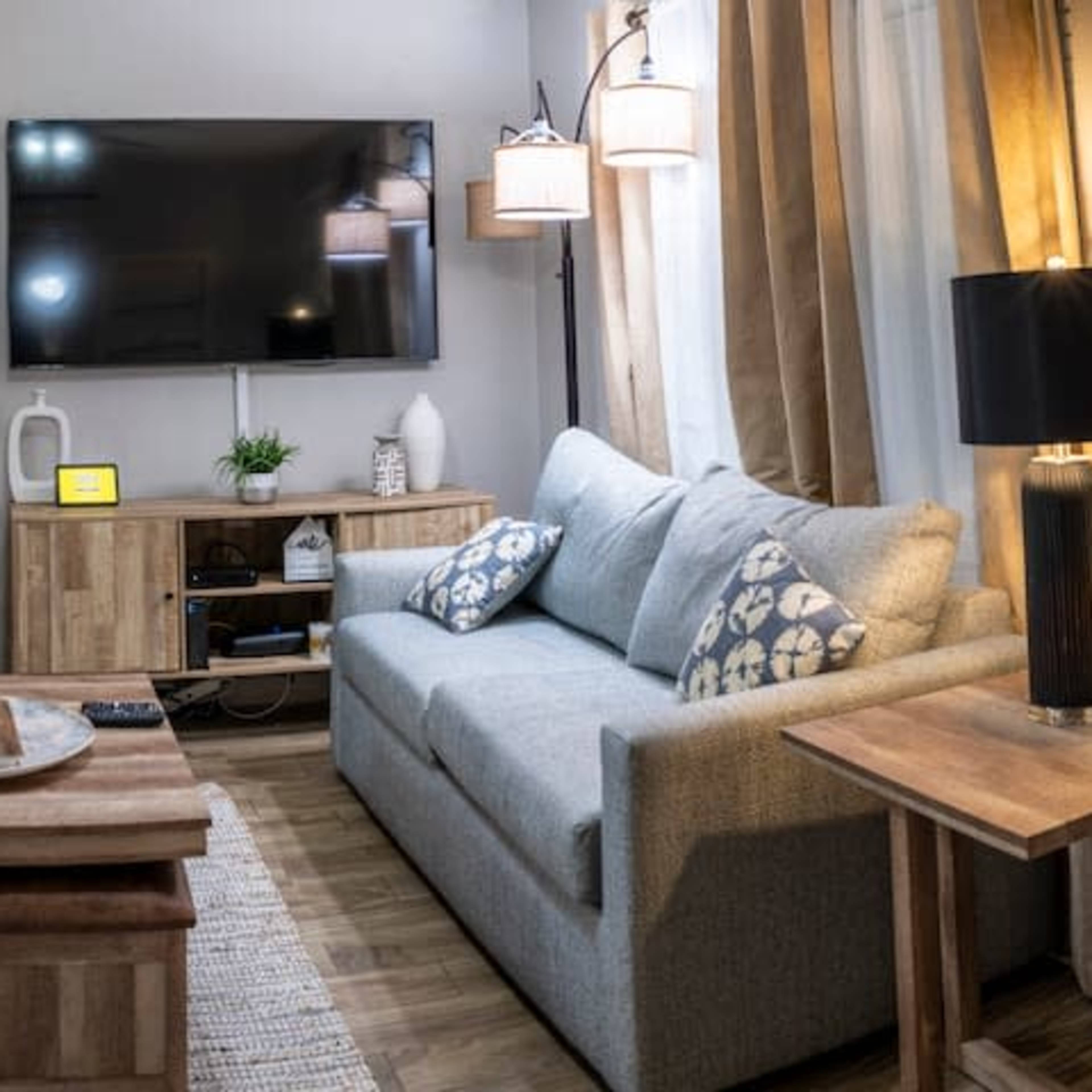 A modern living room with a gray sofa, a wooden coffee table, and a wall-mounted television, adjacent to a small kitchen area with bar stools.