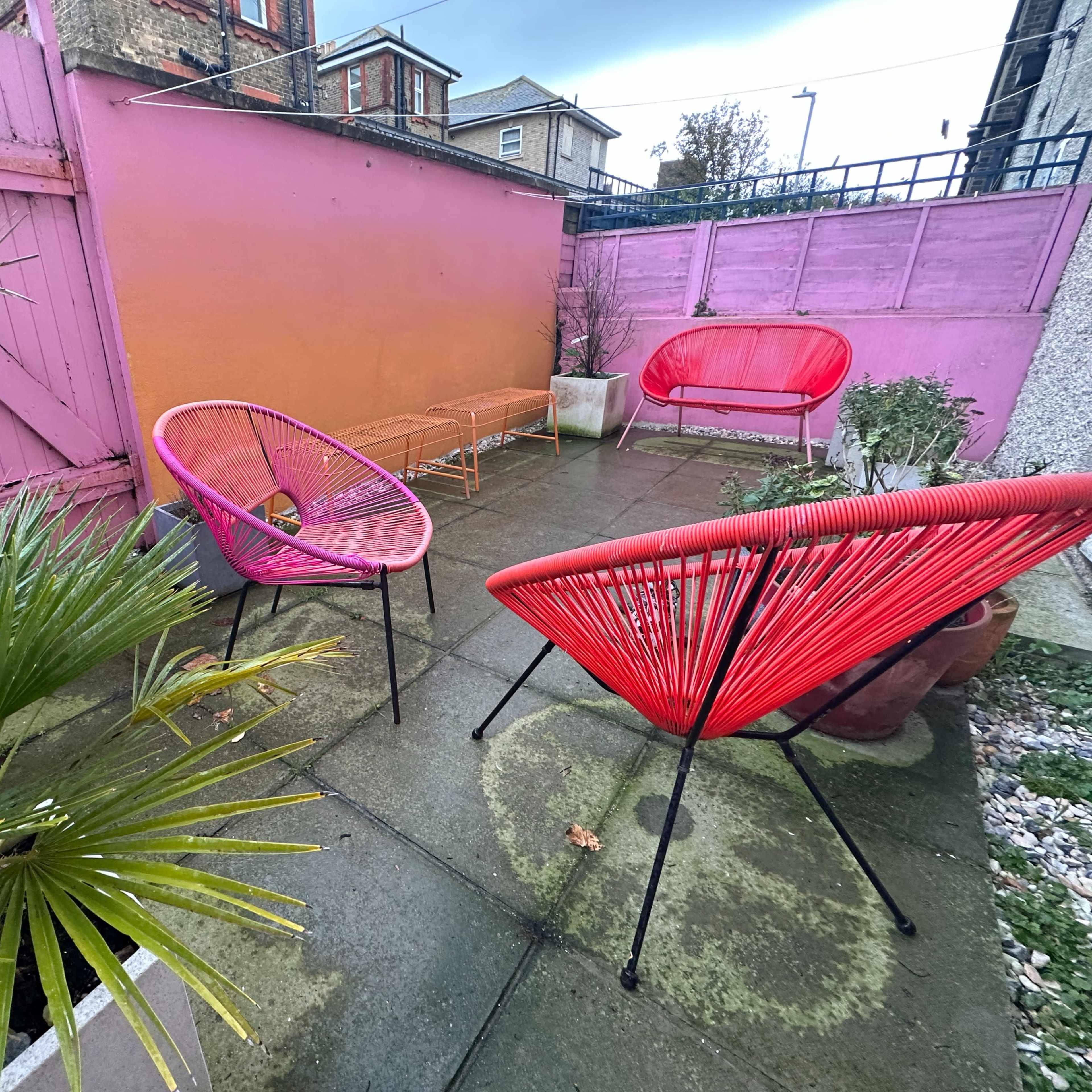 A small outdoor patio features colorful chairs surrounded by greenery and a vibrant pink wall.
