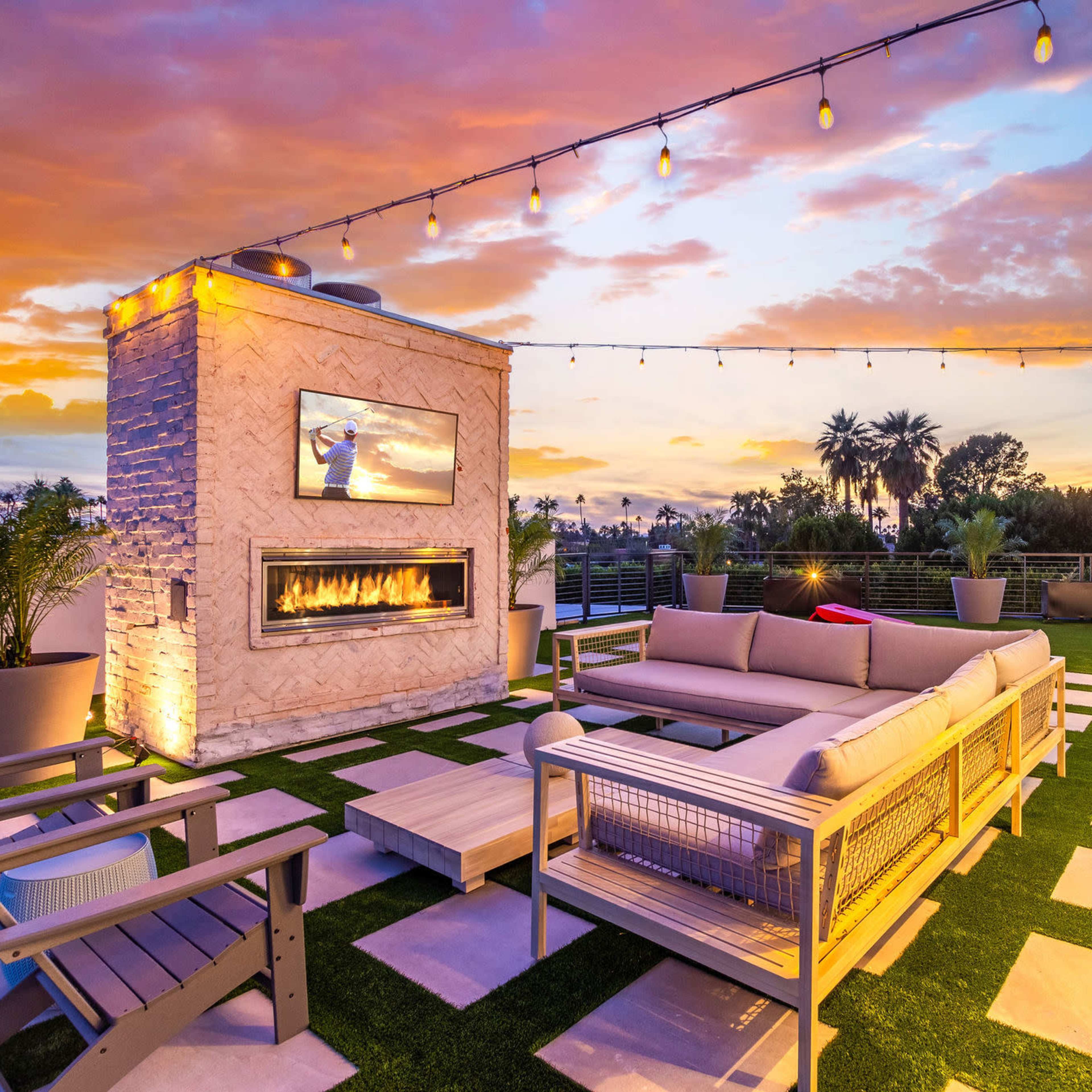 A modern rooftop terrace with a fire pit, a large sofa, and decorative lighting against a vibrant sunset sky.